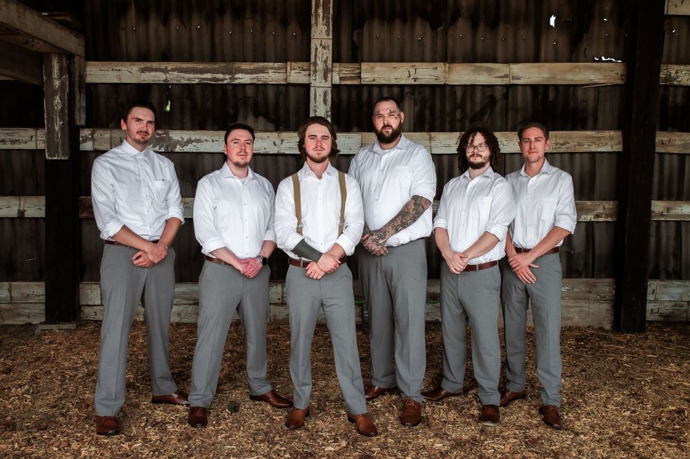 Wedding party members in grey suits sit casually on rustic wooden barn beams.