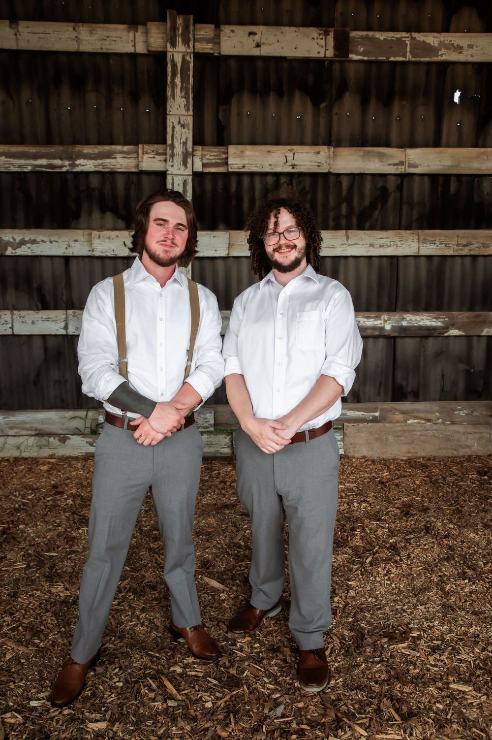 Two people in coordinated vintage outfits stand together on mulch floor inside wooden barn structure.