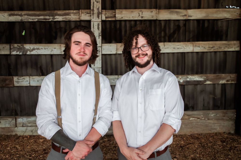 Two people wearing suspenders and dress clothes stand together in rustic barn environment.