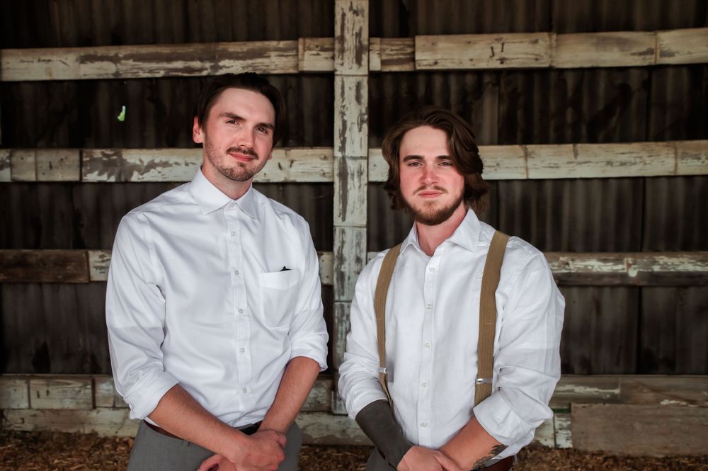 Two people in vintage-style clothing pose casually against wooden barn interior walls.