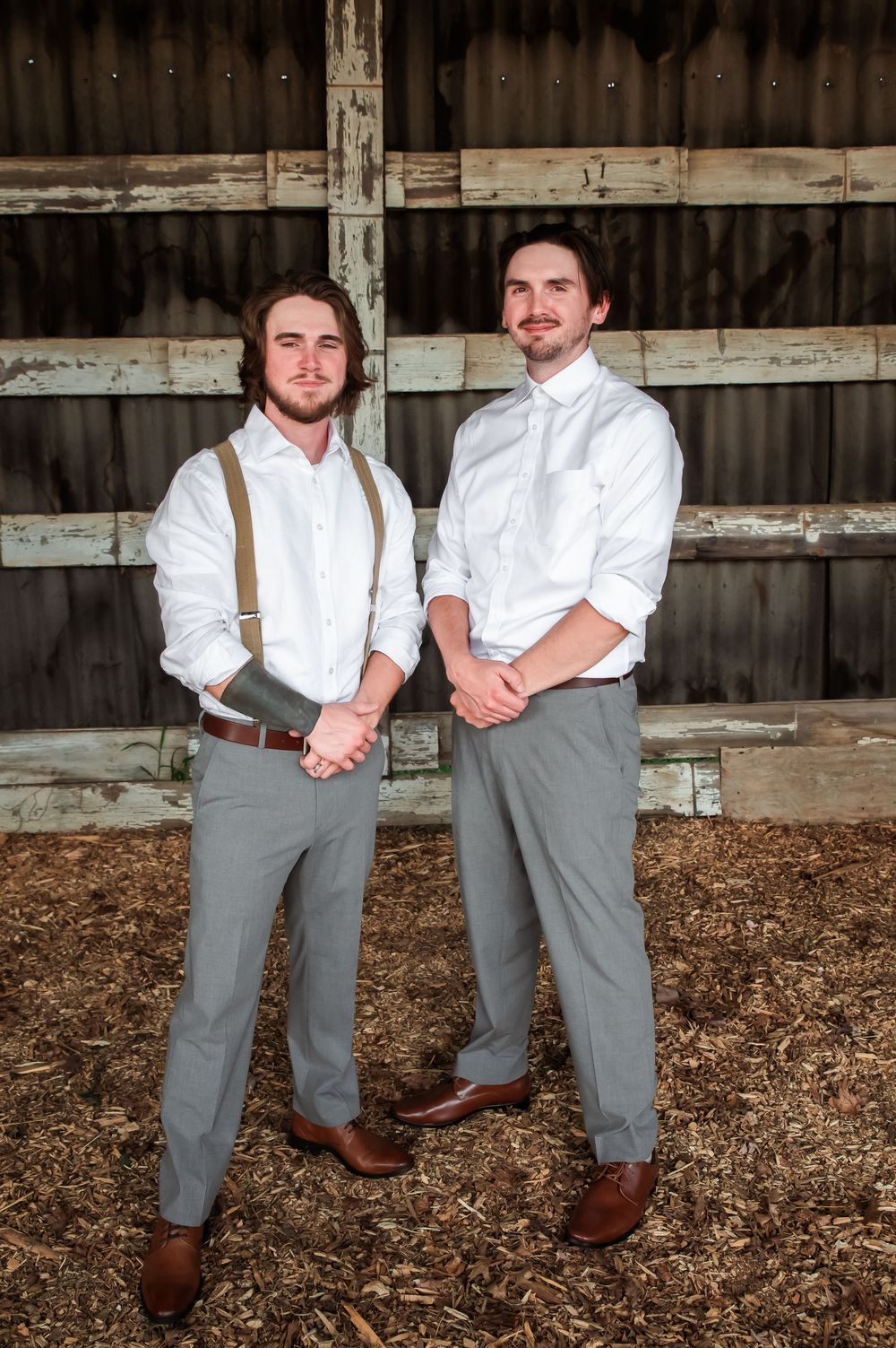 Two people in matching white shirts and gray pants pose in front of weathered barn walls.