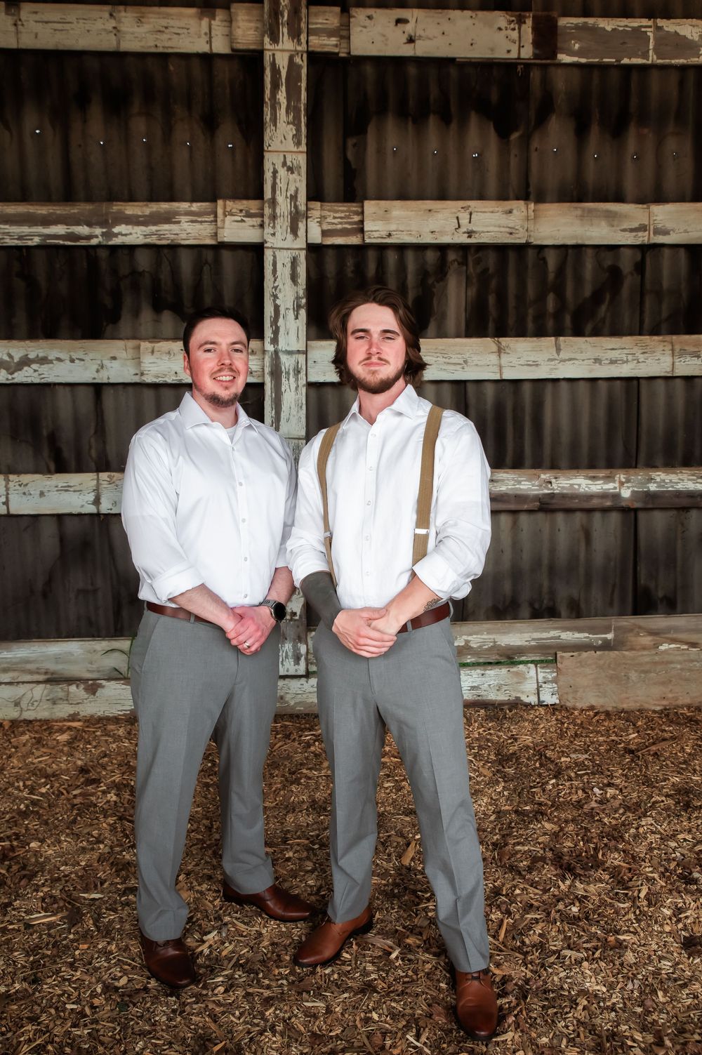 Two people in coordinated formal attire stand together on mulch floor in rustic barn setting.