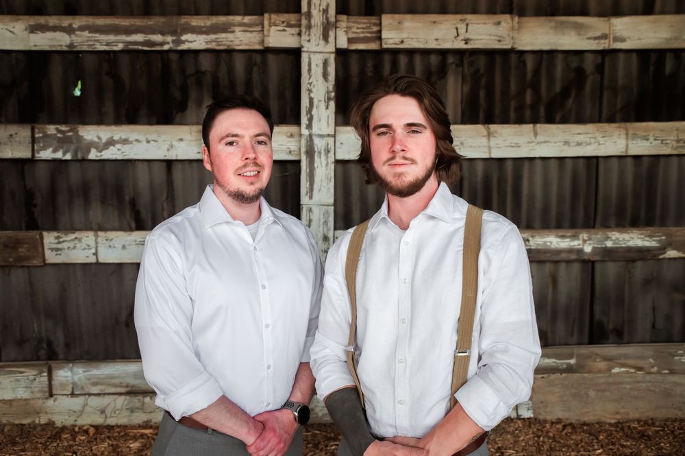 Two people in vintage dress clothes and suspenders stand together inside rustic wooden barn.