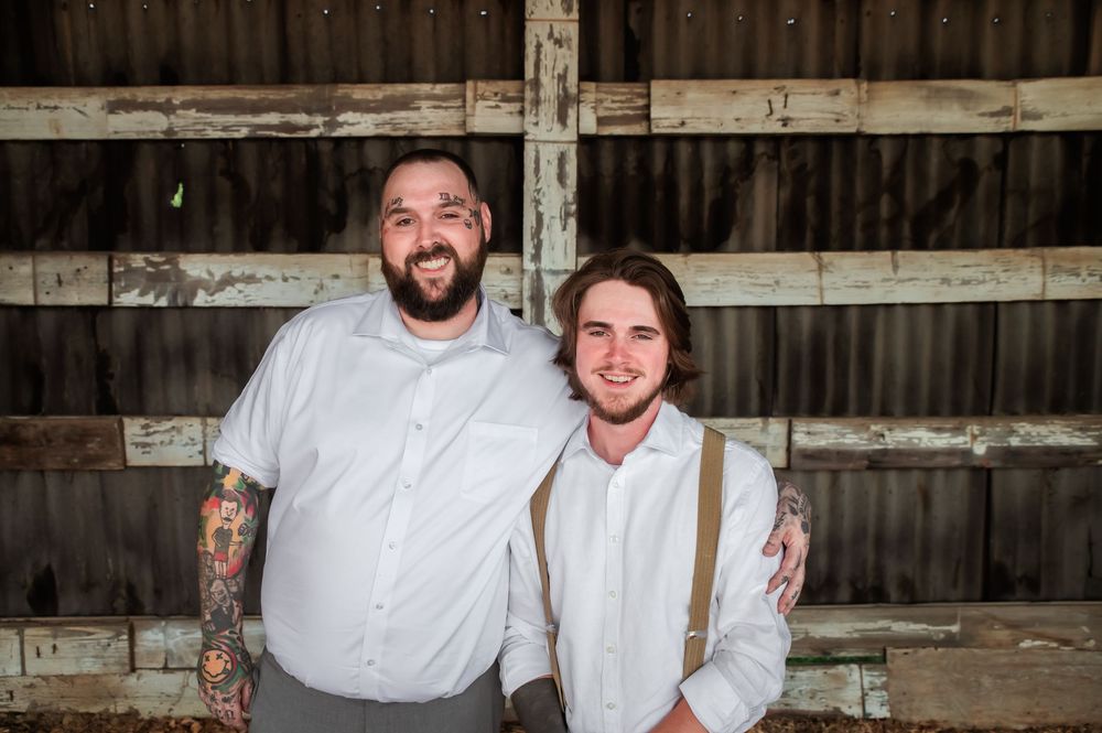 Two people in white shirts and gray pants pose together in a rustic barn setting.