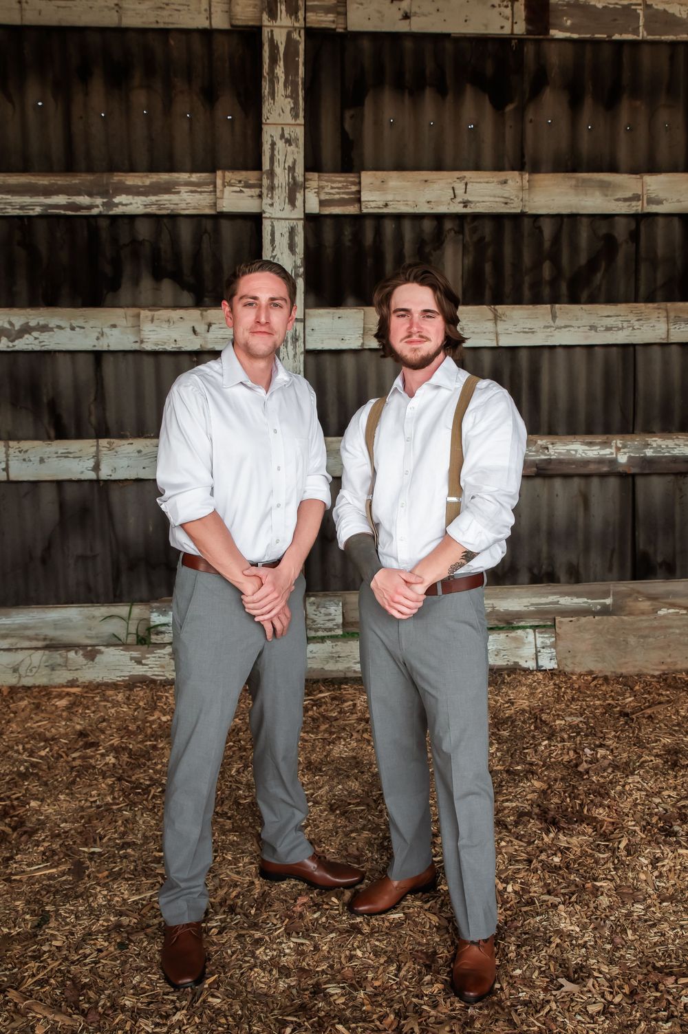Two people in matching white shirts and suspenders sit casually on wooden beams in a barn.