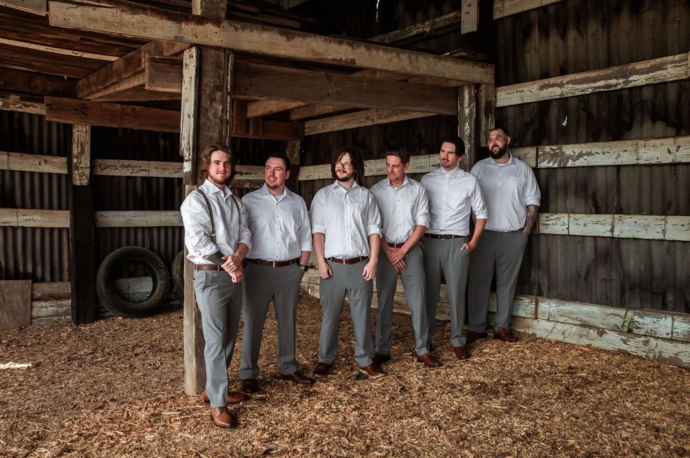 Wedding party lined up against barn wall wearing matching gray pants and white shirts.