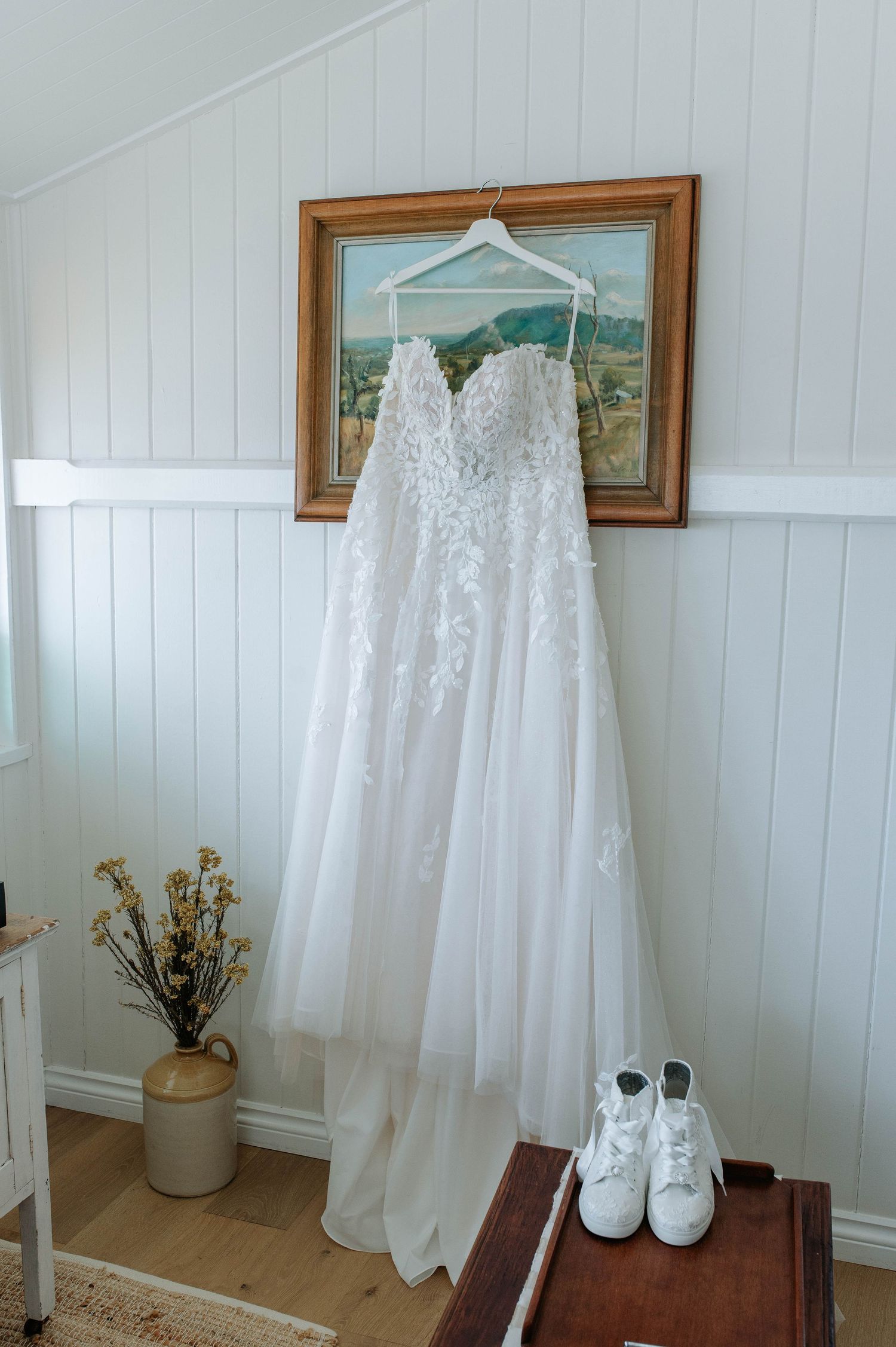 A white lace wedding dress hangs on a hanger against a wall with white wainscoting, paired with white boots below.