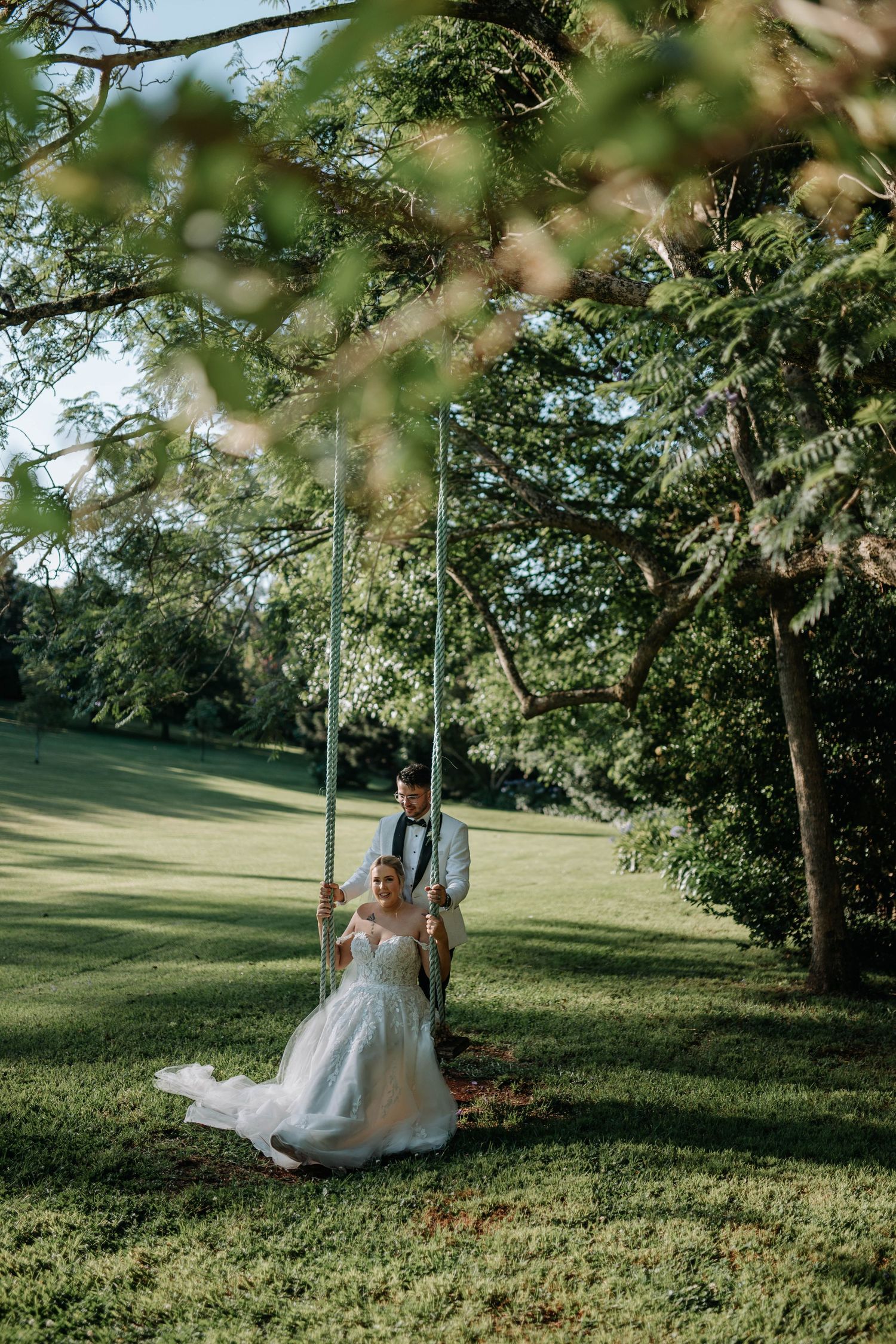 A couple in wedding attire share a romantic moment on a tree swing in a sunlit garden setting.