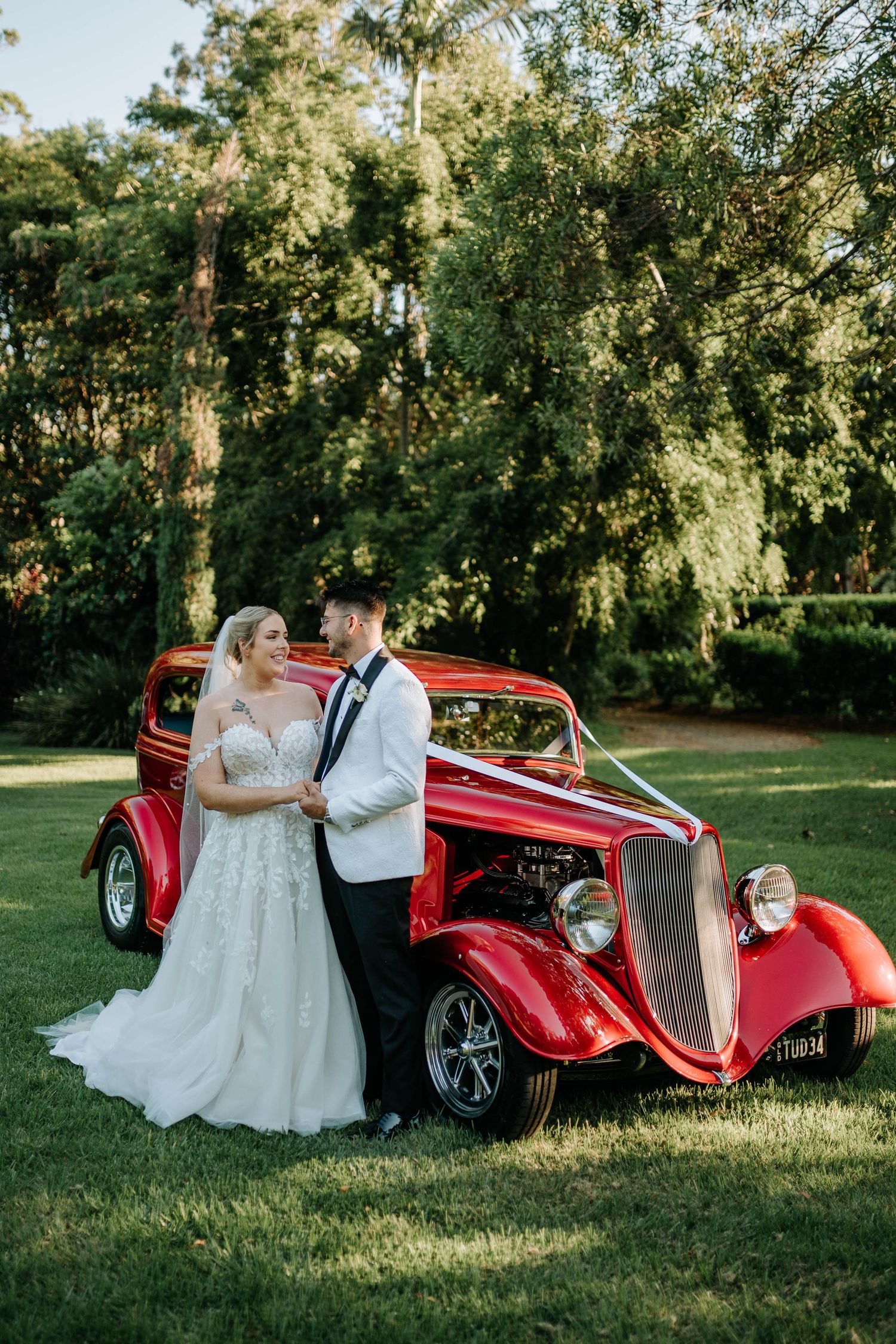 A vintage red hot rod car serves as a romantic backdrop for a wedding photo session in a sunlit outdoor setting.