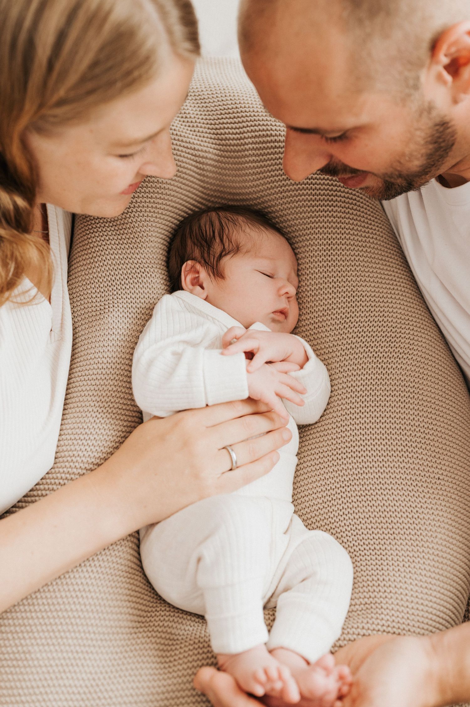 A sleeping baby in white clothing rests peacefully on a beige textured surface while being gently held.