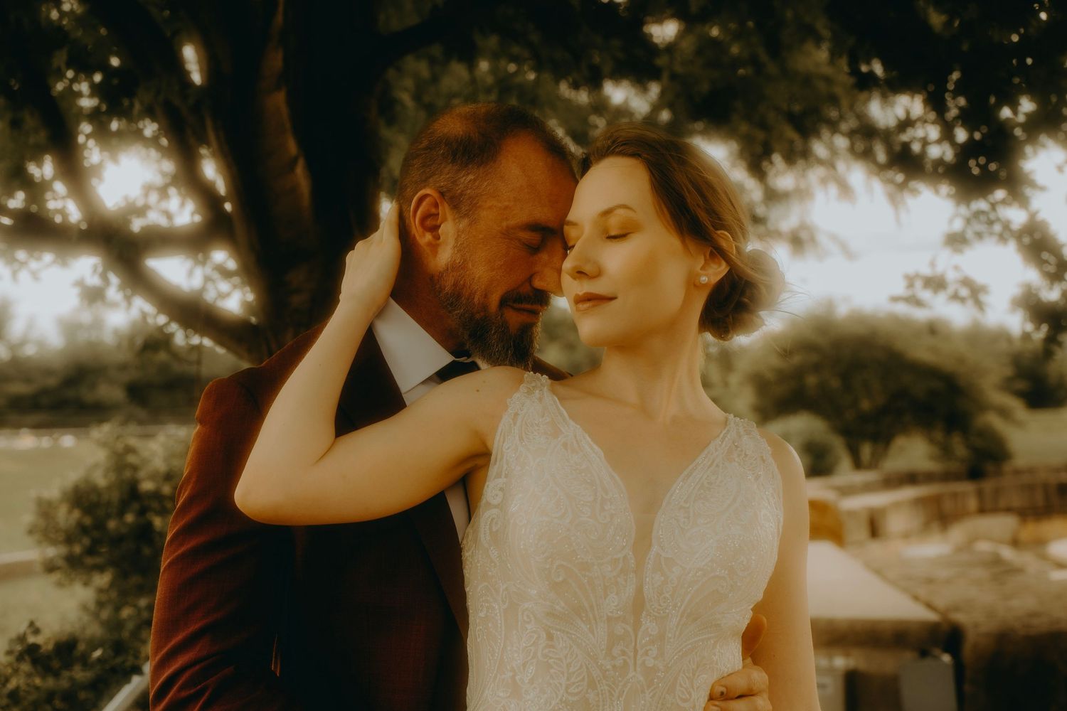 Romantic wedding couple embracing outdoors, bride in white gown with groom in burgundy suit under a tree.