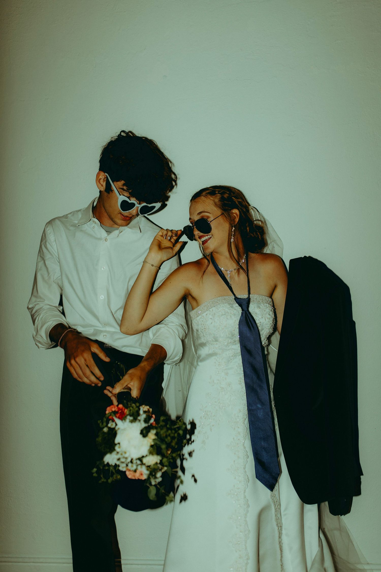 Bride and groom in sunglasses posing playfully with wedding bouquet, bride wearing grooms tie and jacket.