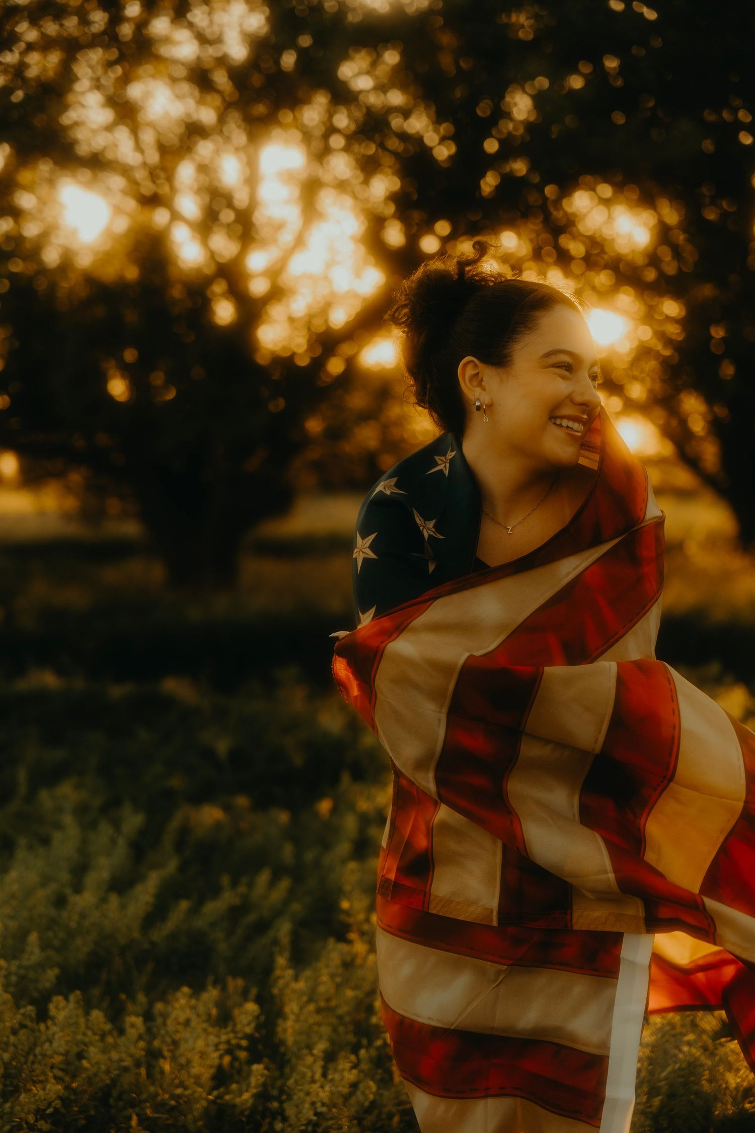 Woman wrapped in American flag smiling at golden sunset in a lush green field.