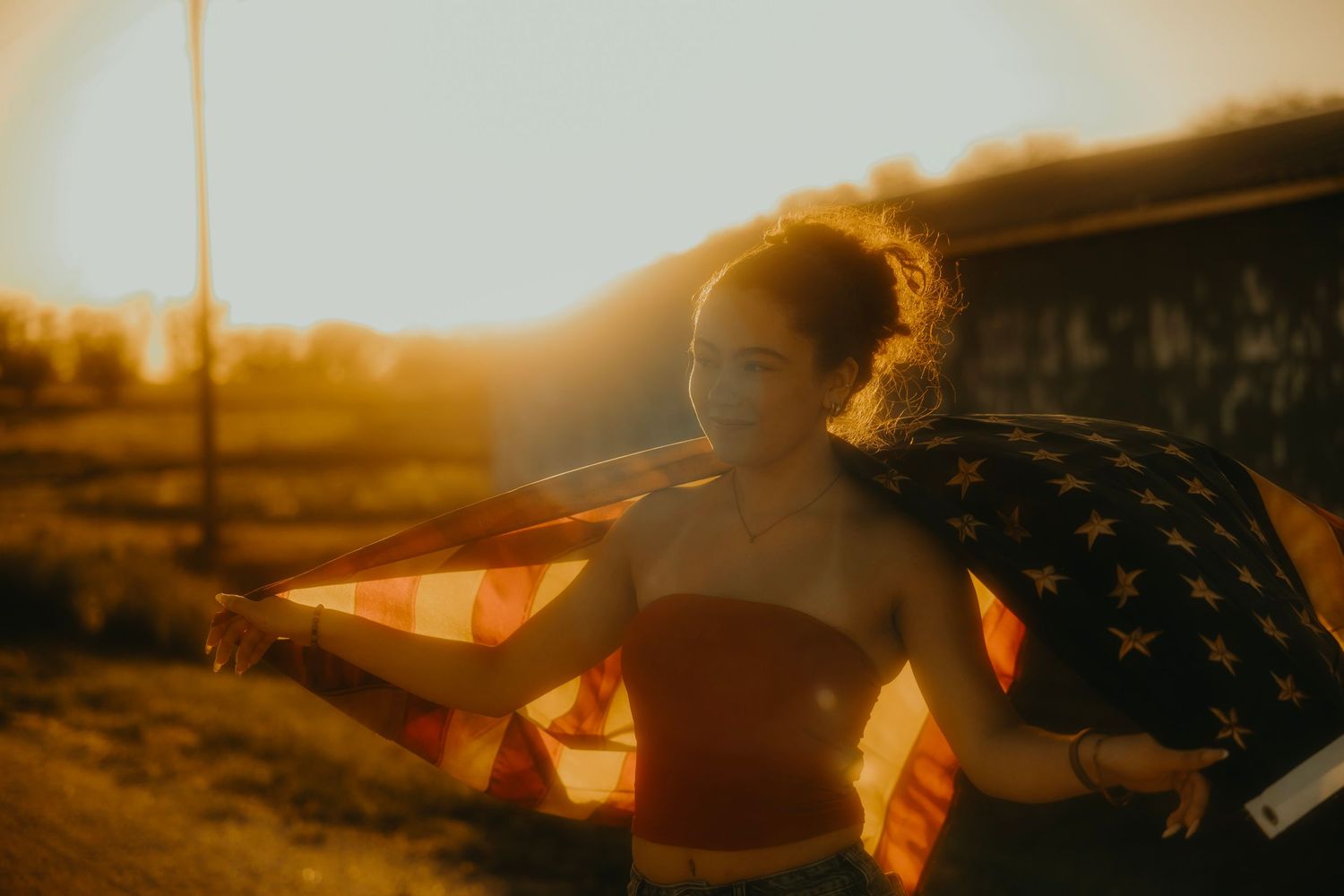 Young woman wearing a face mask draped in an American flag at golden hour sunset outdoors.