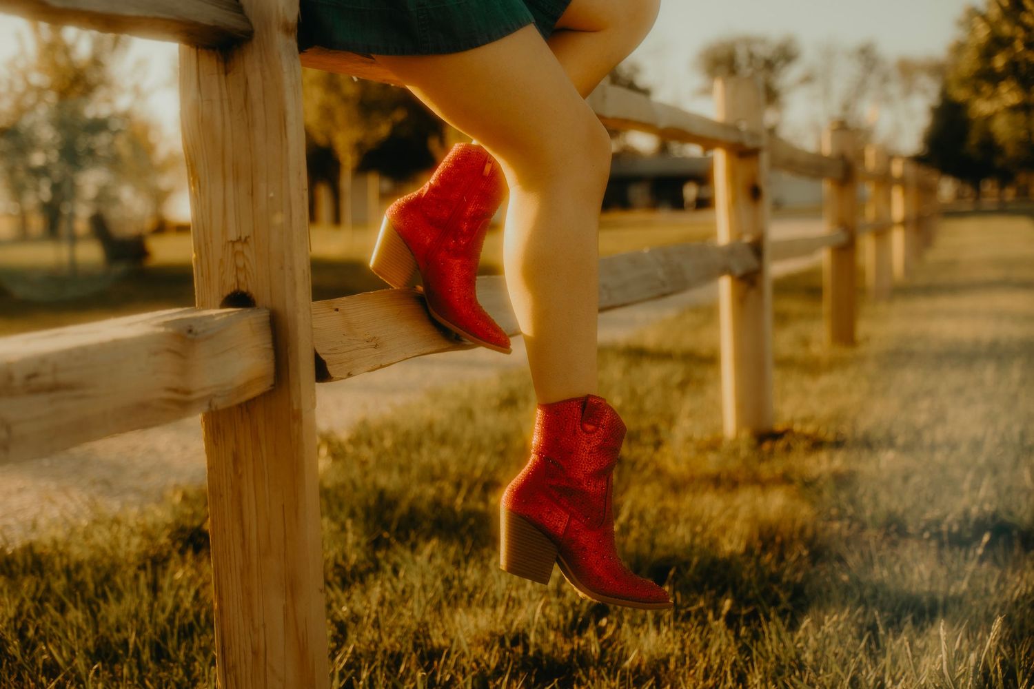 Woman wearing red cowboy boots sitting on a wooden fence rail in golden sunset light outdoors.