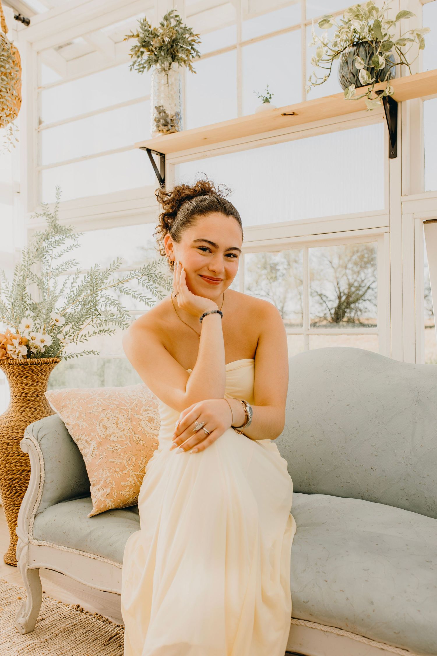 Smiling bride in strapless ivory gown seated on vintage blue sofa in bright sunlit greenhouse setting.