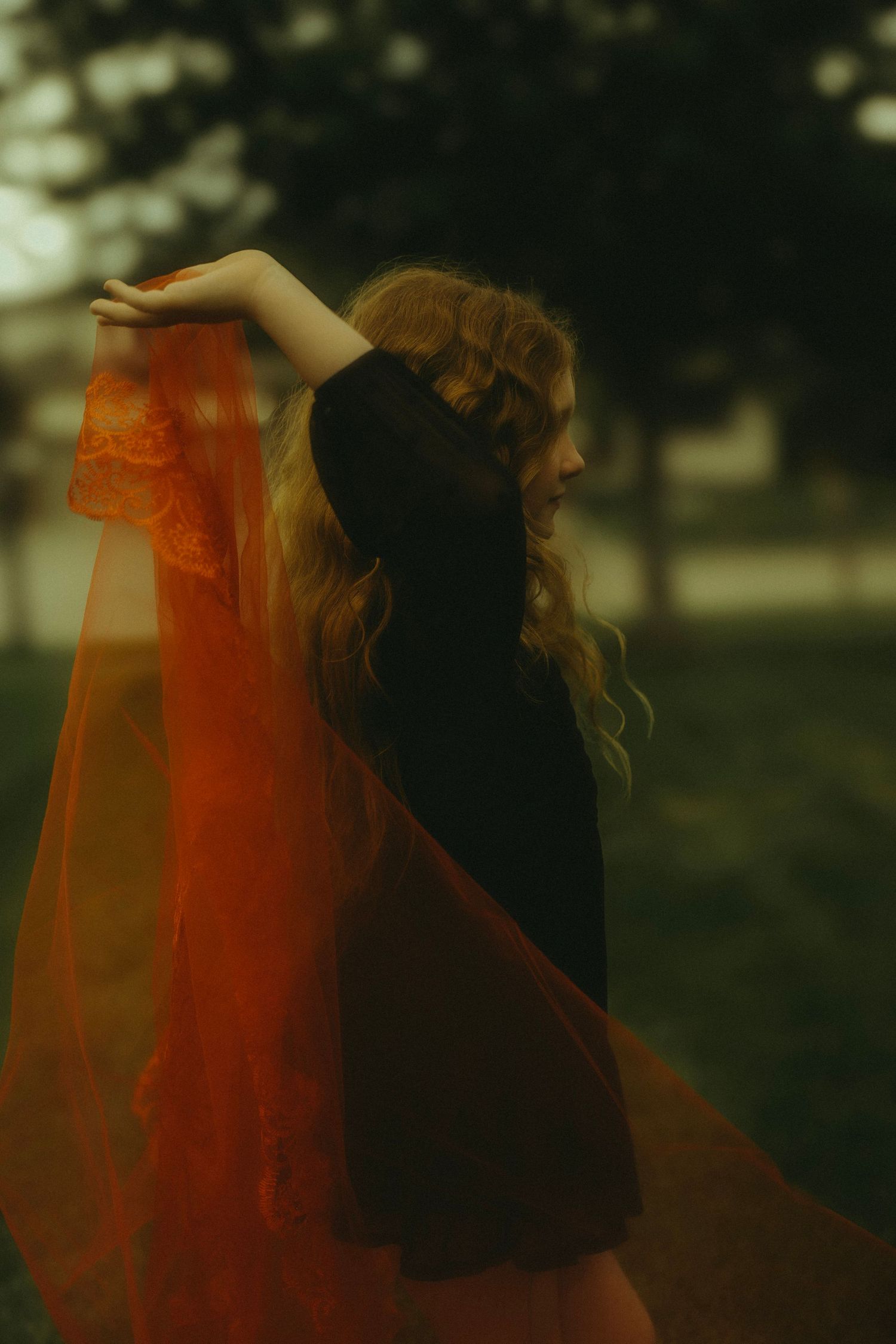 Woman in black dress holding flowing red orange lace fabric outdoors in moody evening light.