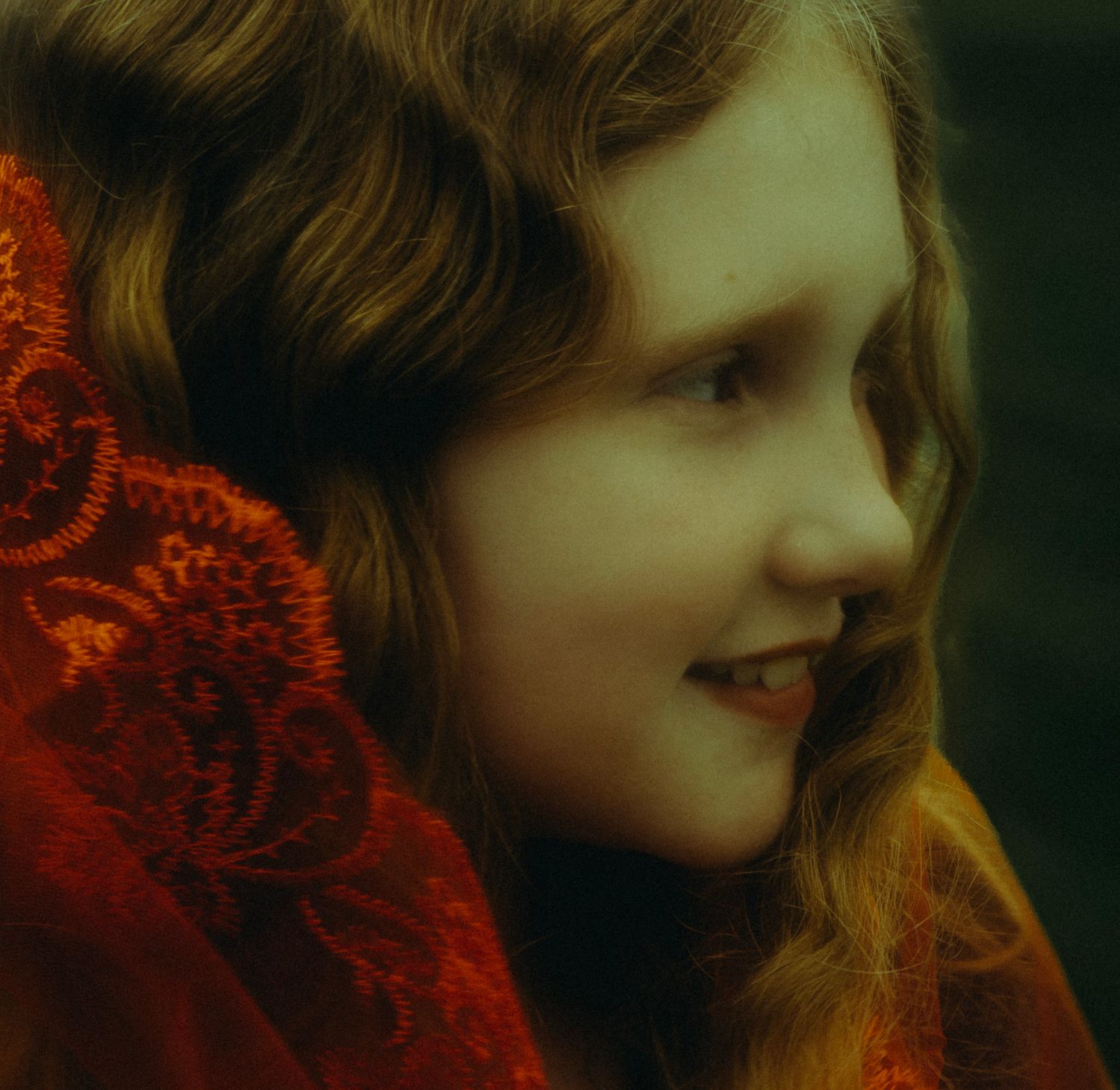 Young girl with wavy auburn hair wearing a red lace shawl, smiling softly in a warm, moody portrait.