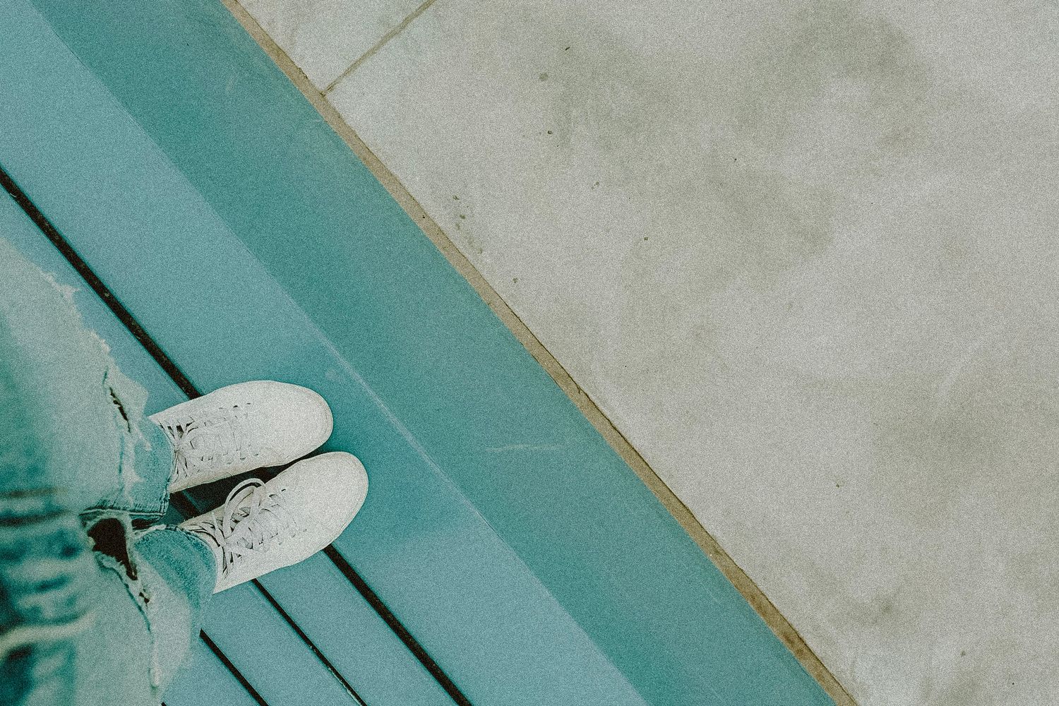 Person standing at the edge of a turquoise pool in white sneakers and light blue jeans on a concrete surface.