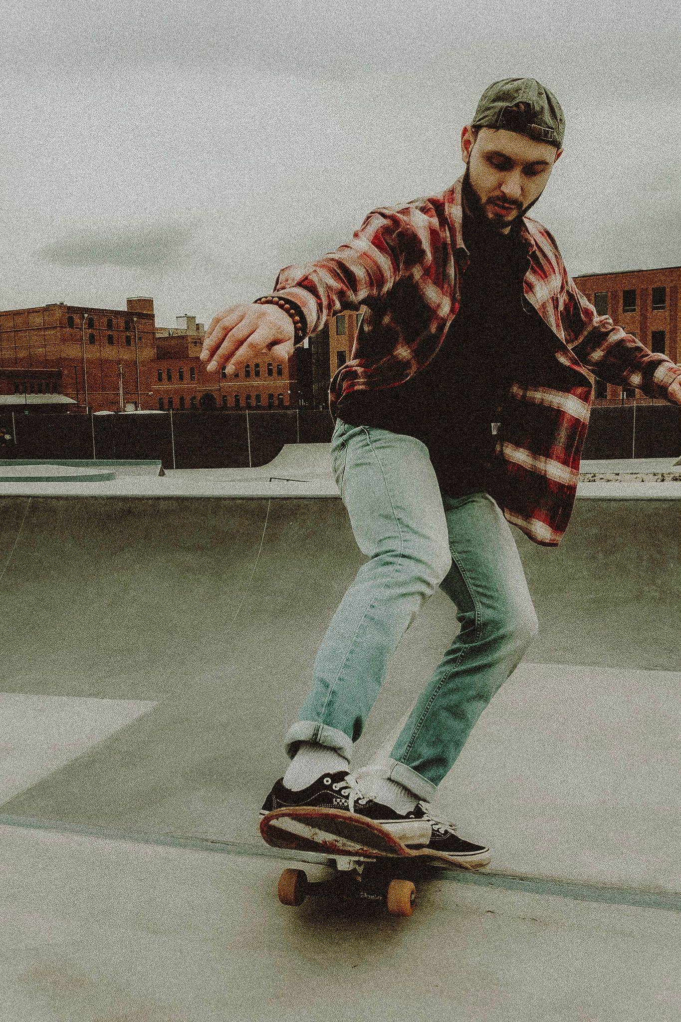A bearded skateboarder in a red flannel shirt and jeans performs a trick at an outdoor skate park.