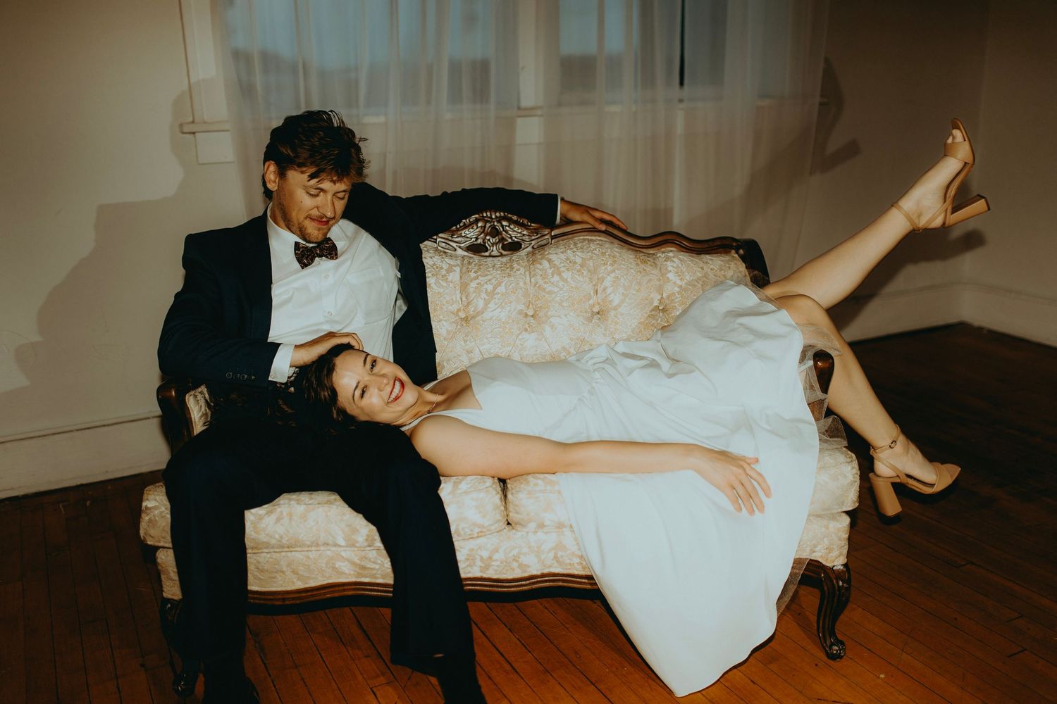 Bride in white gown reclines on vintage sofa beside groom in black tuxedo during elegant wedding portrait session.