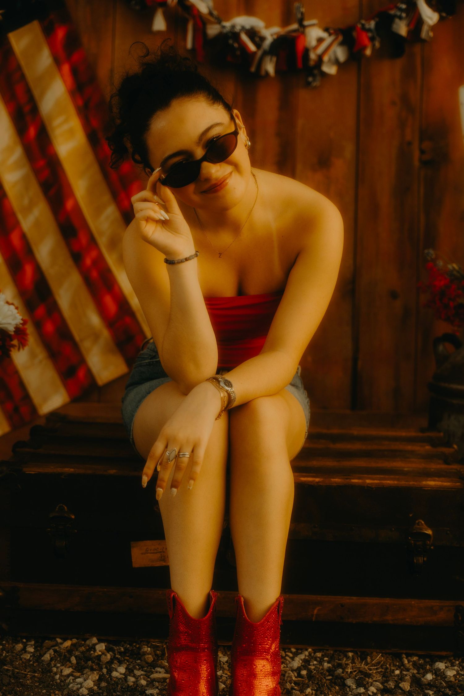 Stylish woman in red tube top, denim shorts, and red cowboy boots posing with sunglasses near American flag.