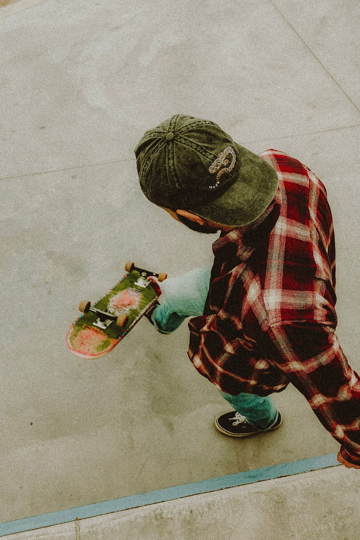 Skateboarder in red plaid shirt and green beanie crouching at skate park, holding a skateboard.
