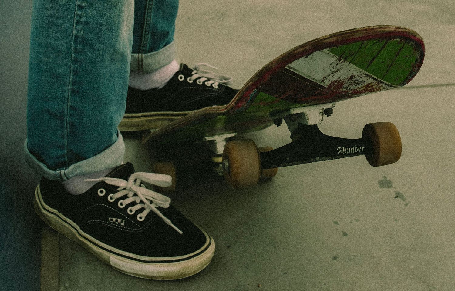 Person wearing black Vans sneakers standing on a green and red skateboard on pavement.