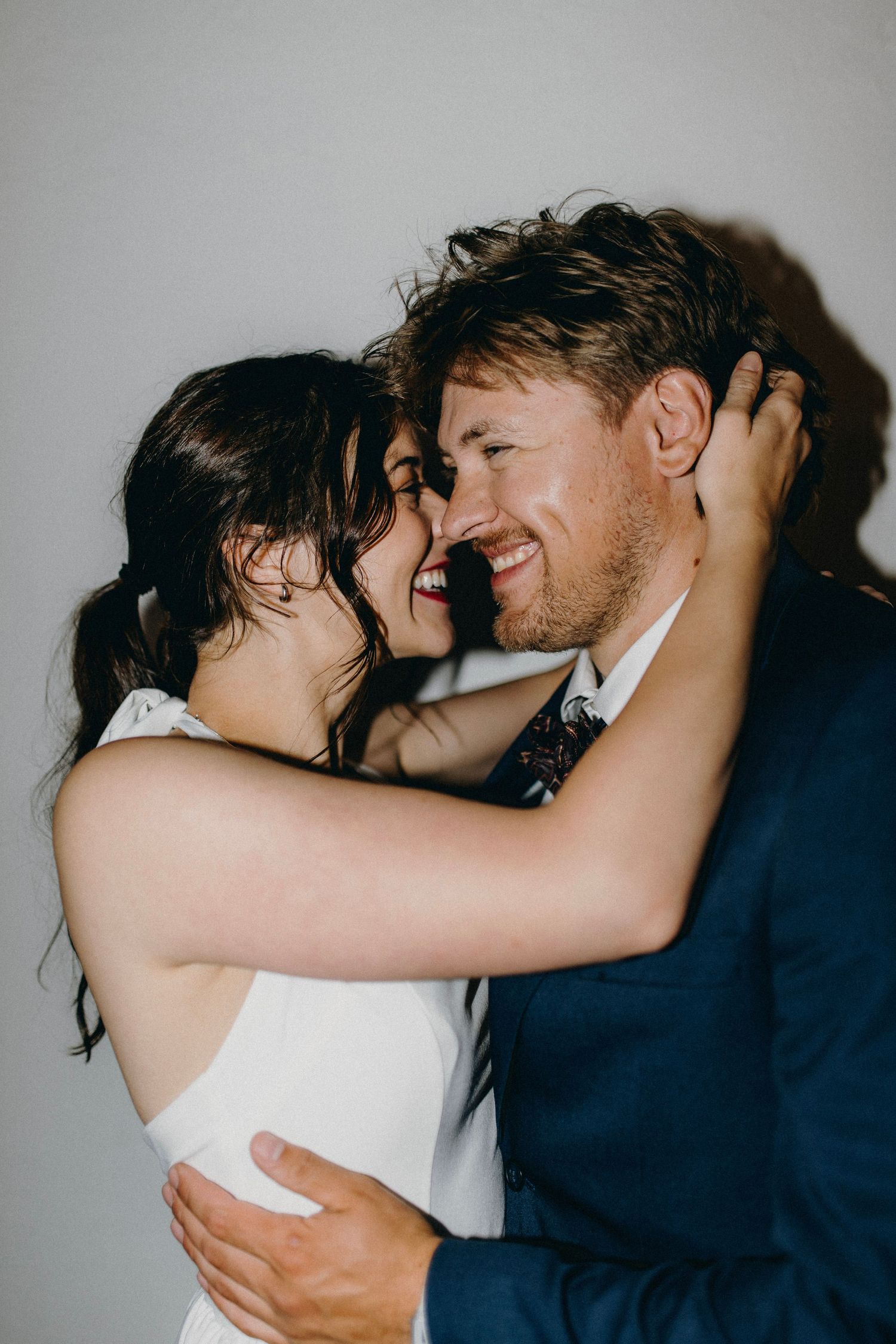 A smiling groom in a navy suit embraces his bride in a white dress as they share a romantic moment.