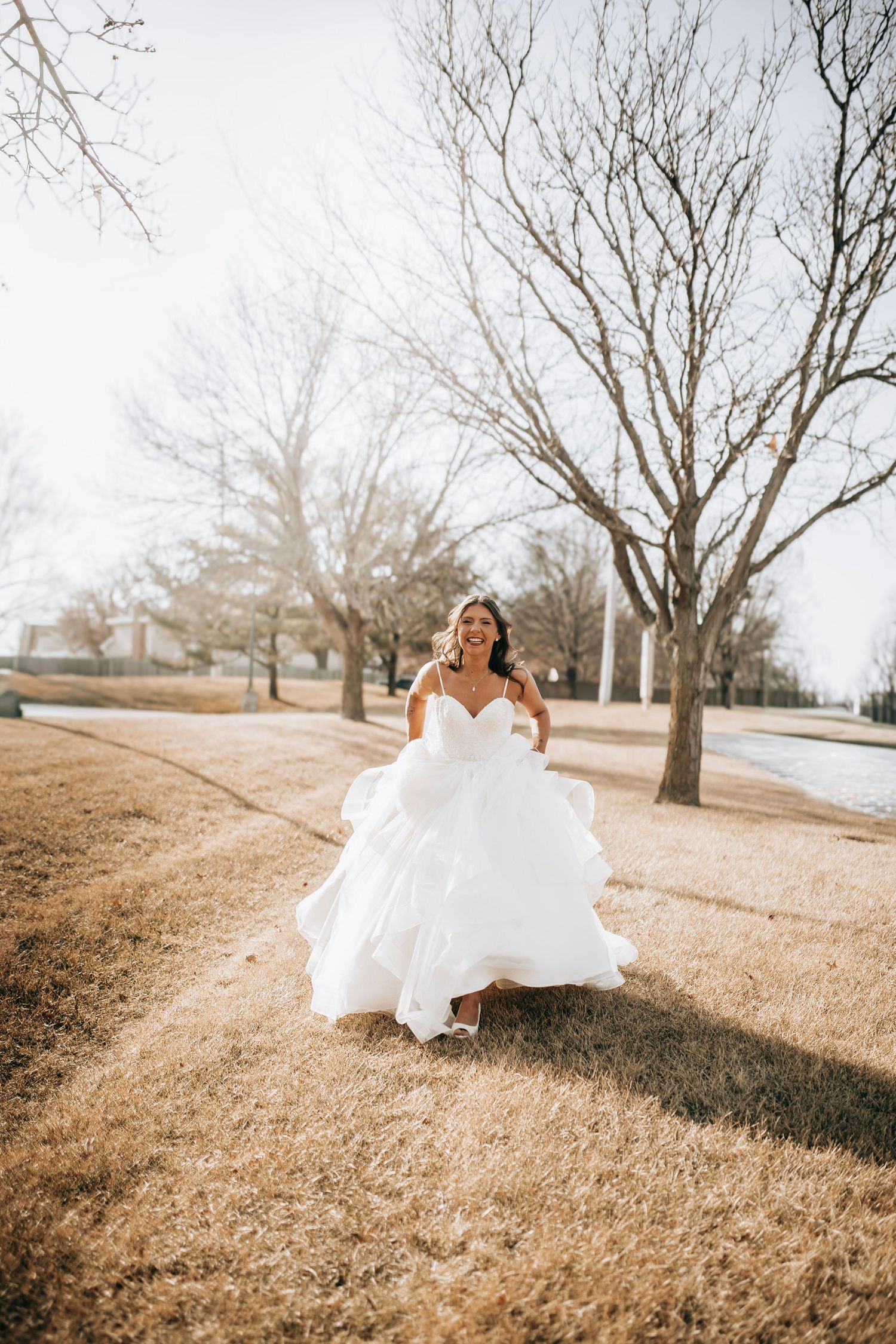 A bride in a flowing white gown twirls joyfully on a grassy hillside during a golden hour photo session.