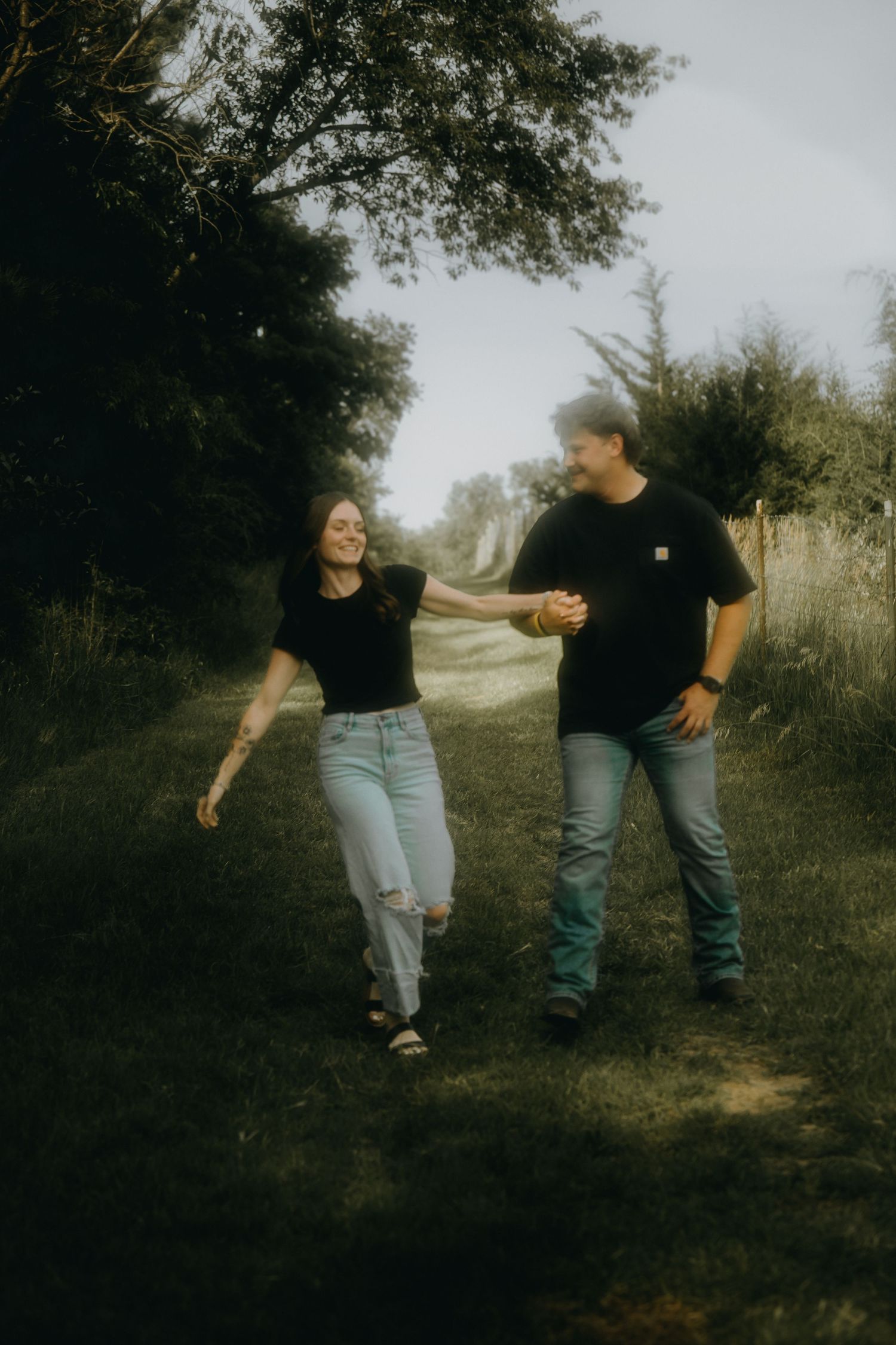 A couple holding hands and walking along a tree-lined path, both wearing black shirts and jeans, smiling at each other.