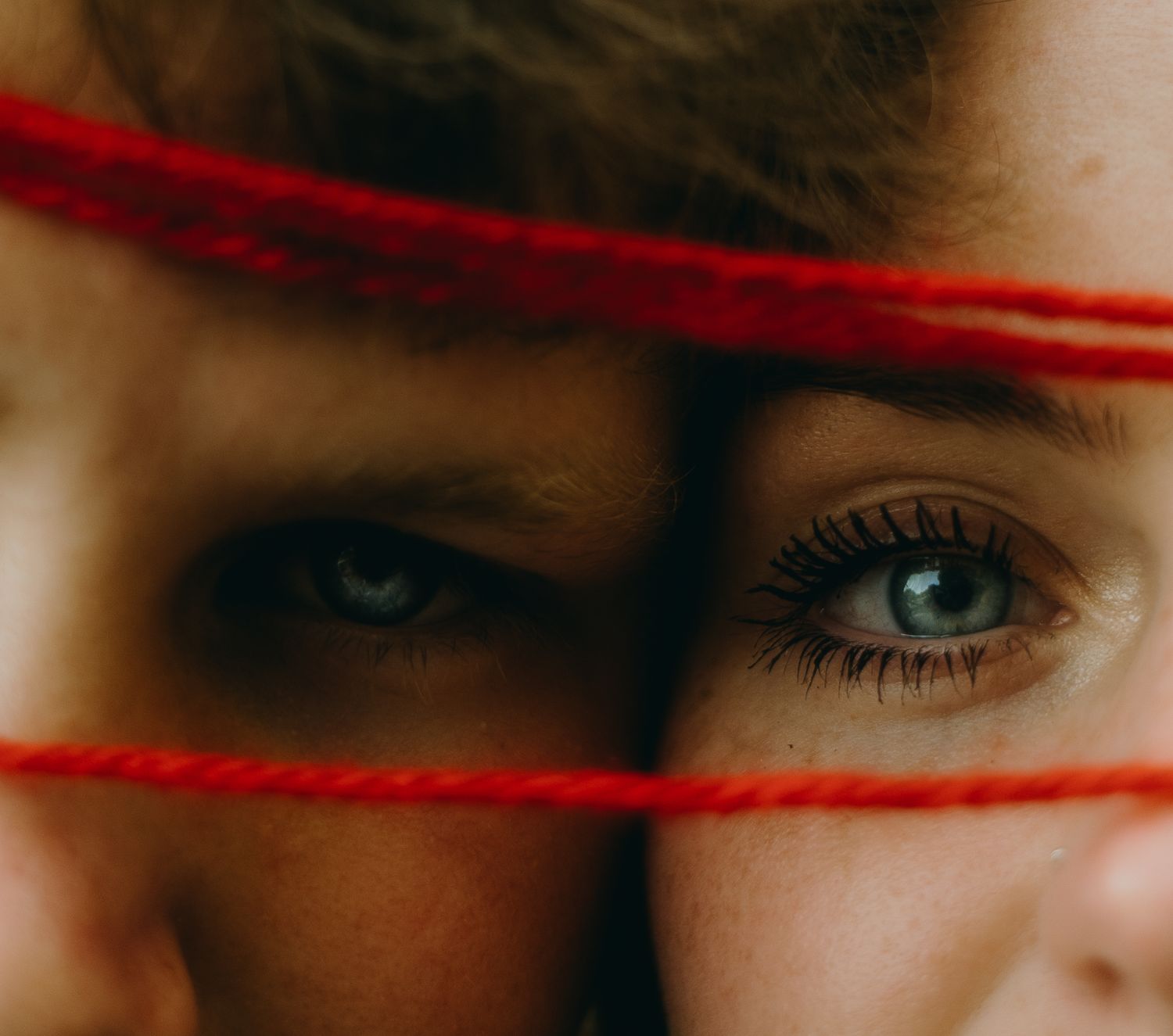 Close-up of two people's eyes partially obscured by red yarn strings in an artistic portrait photo.