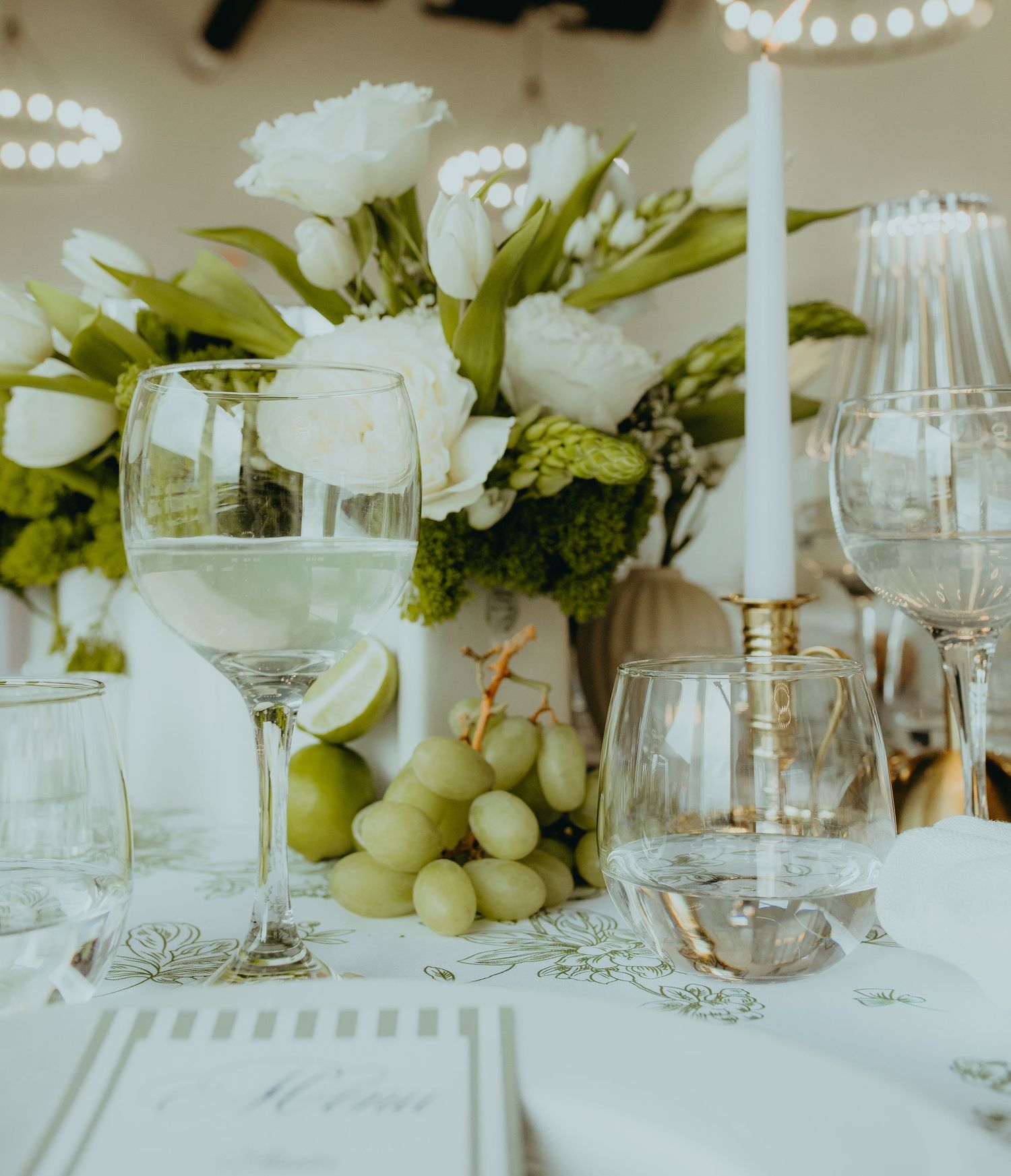Elegant wedding table setting with white roses, green grapes, crystal glasses, and a white taper candle centerpiece.