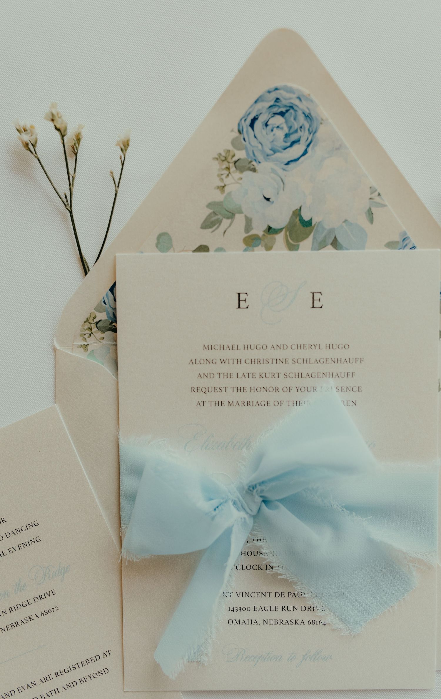 Elegant wedding invitation suite with light blue bow, floral envelope liner featuring blue roses, and classic monogram on cream cardstock.