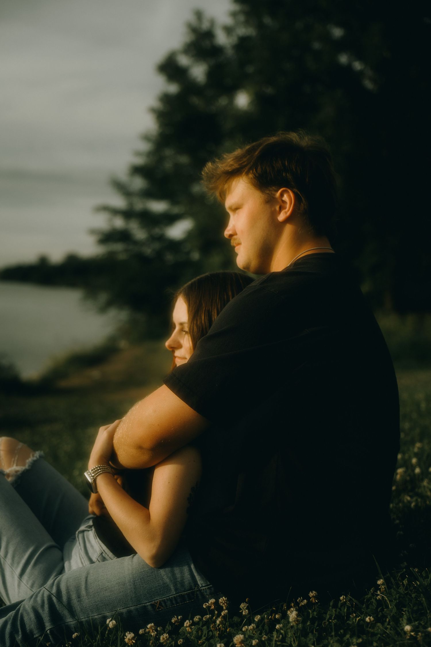 A couple sits embraced on grass at golden hour, with trees and water in the background.