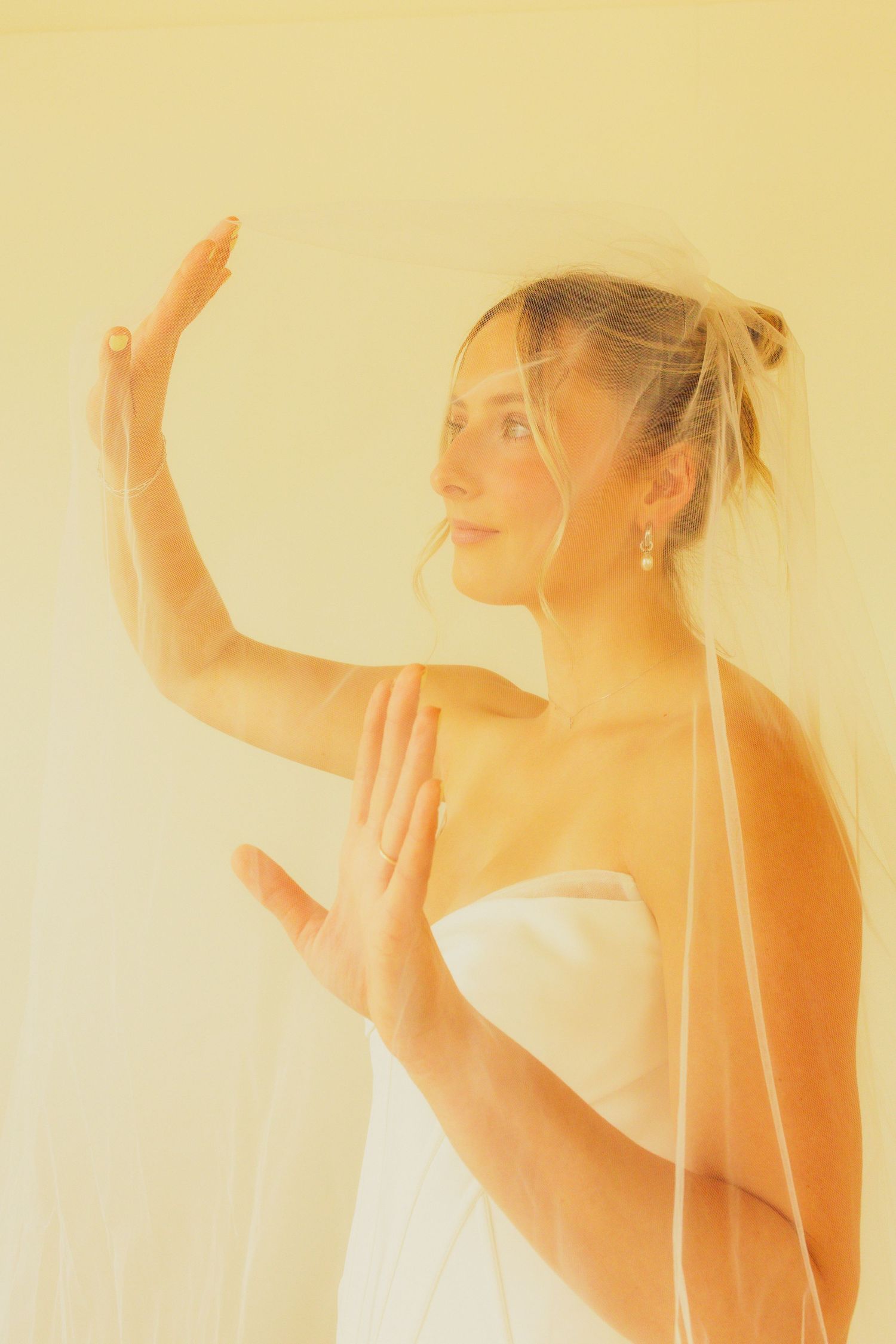 Elegant bride in white strapless gown and veil poses gracefully with raised hands in soft golden light.