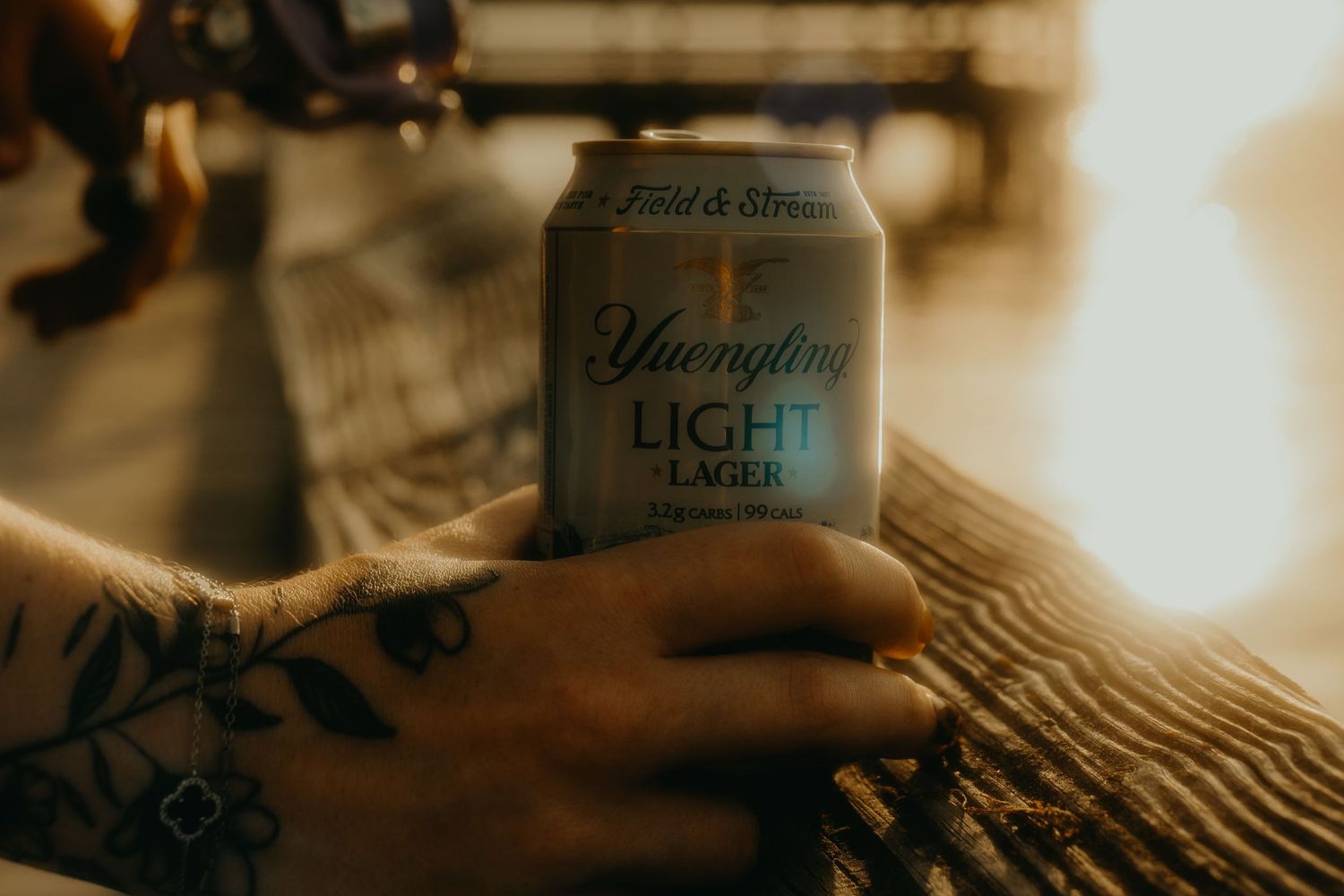 Tattooed hand holding a Yuengling Light Lager beer can in warm golden sunset light.