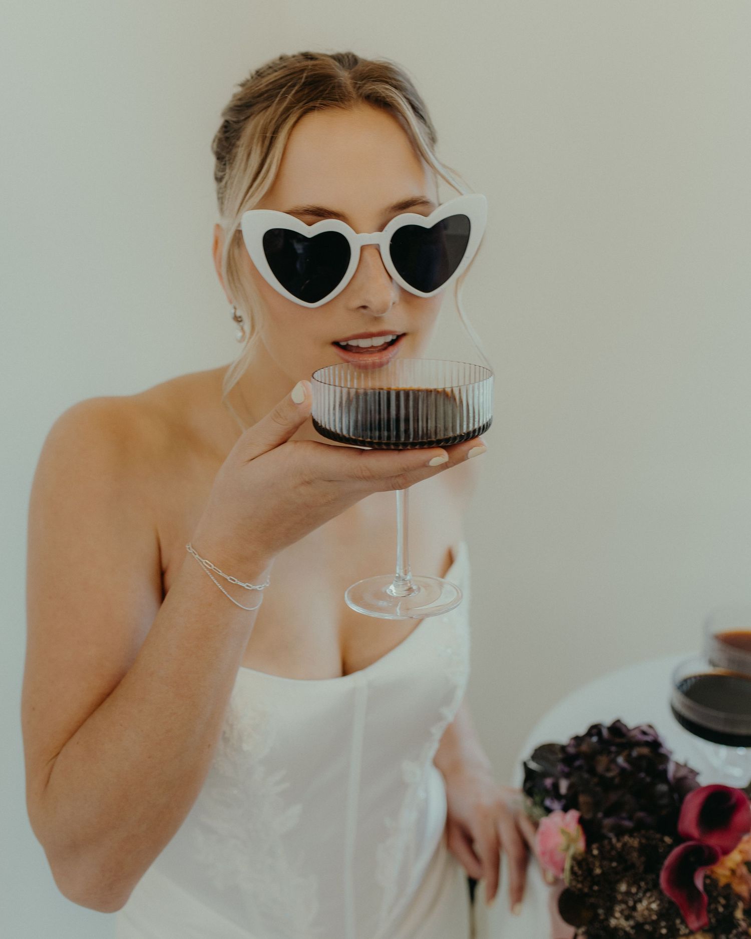 Bride in white dress and heart-shaped sunglasses holding a glass of red wine, smiling elegantly.