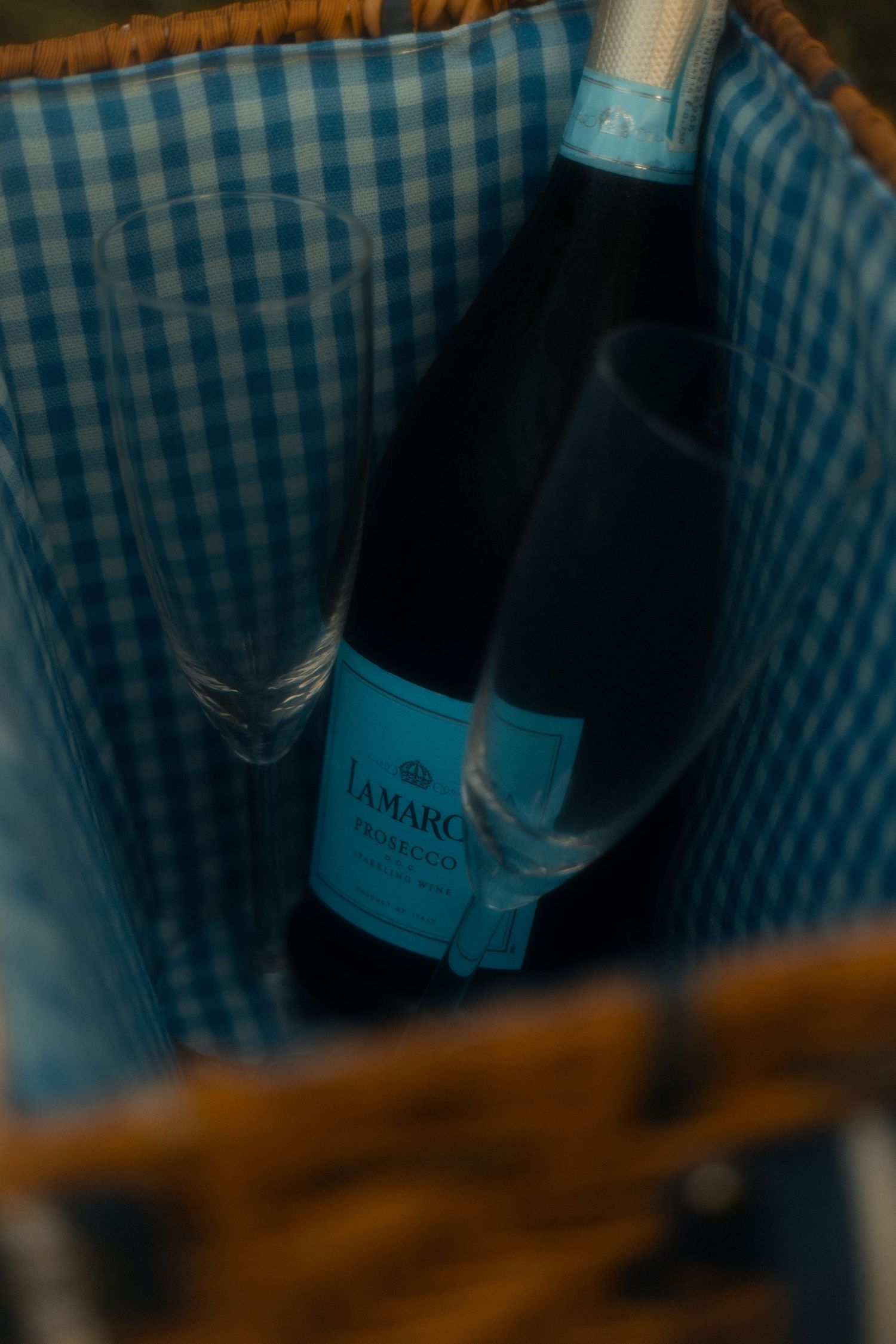 La Marco Prosecco bottle and wine glasses inside a blue gingham-lined wicker picnic basket.
