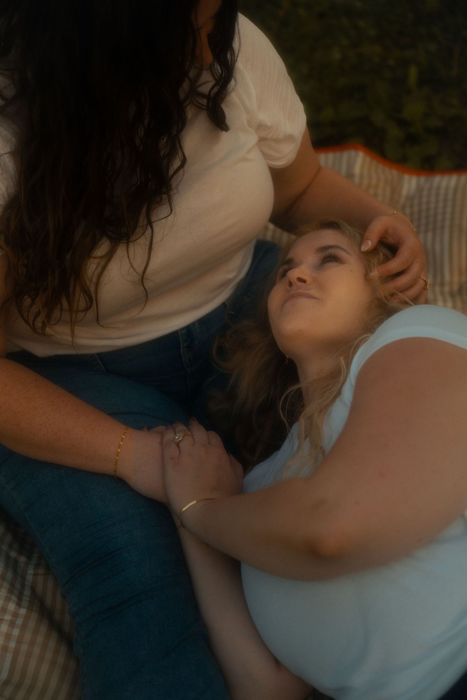 Two women sharing a tender moment outdoors, one lying down looking up while the other gently touches her hair.