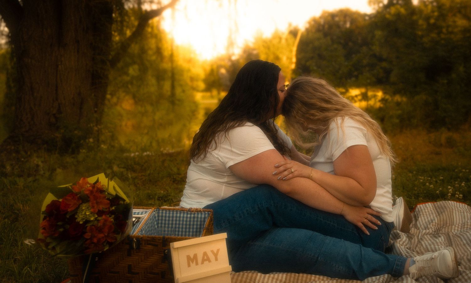 Two women sharing an intimate moment on a picnic blanket outdoors, with golden sunset light filtering through trees.