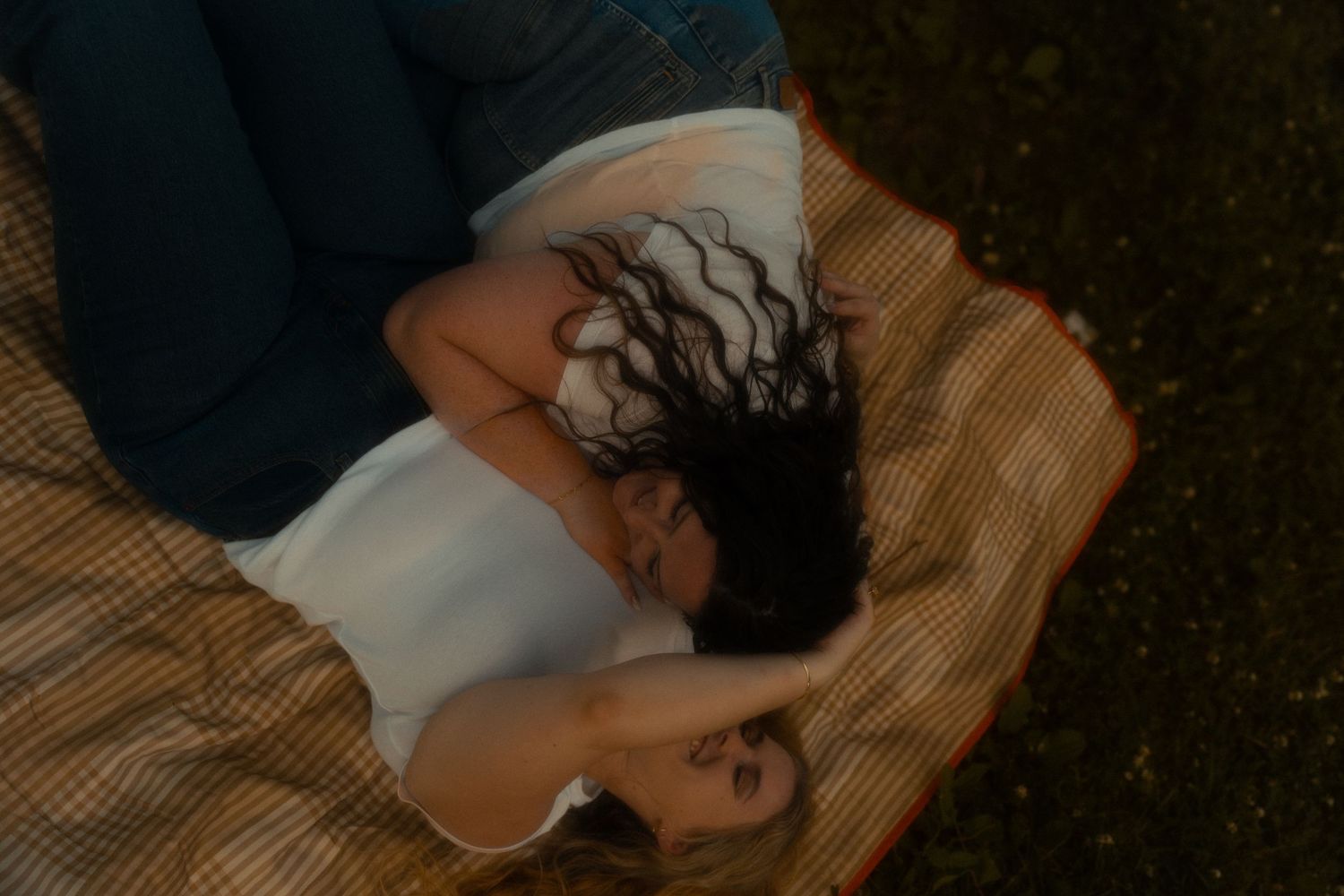 Two women lying on a plaid blanket on grass, viewed from above, in warm golden light.