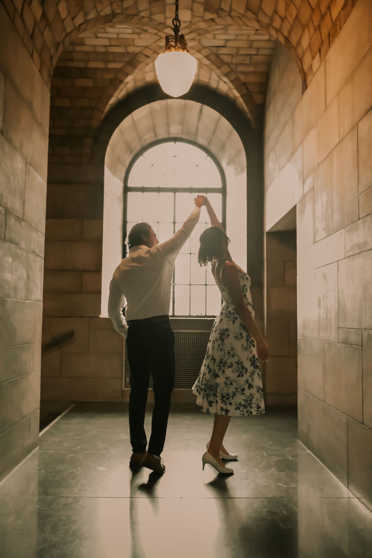 Two people dance gracefully in a sunlit hallway with arched stone walls and dramatic lighting.