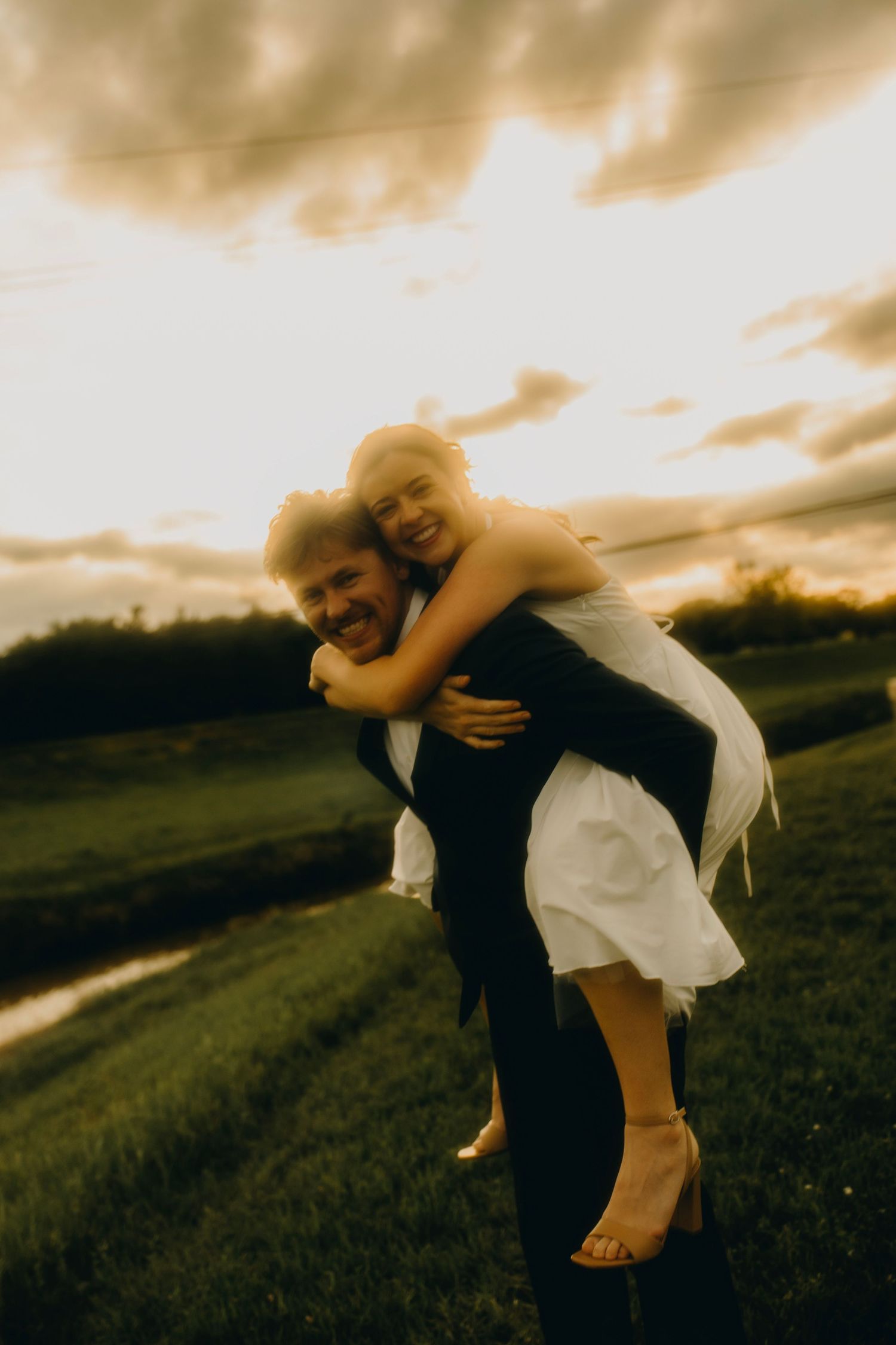 Groom giving bride a piggyback ride in a green field at golden sunset, both laughing joyfully in wedding attire.