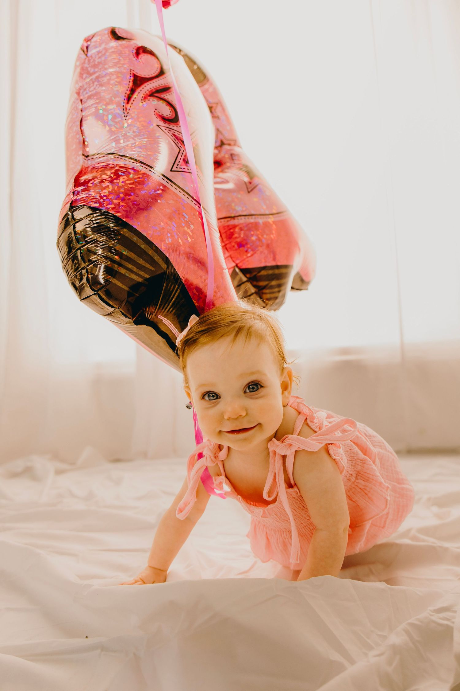 Cute baby girl in pink dress crawling on white bed under a flamingo balloon.