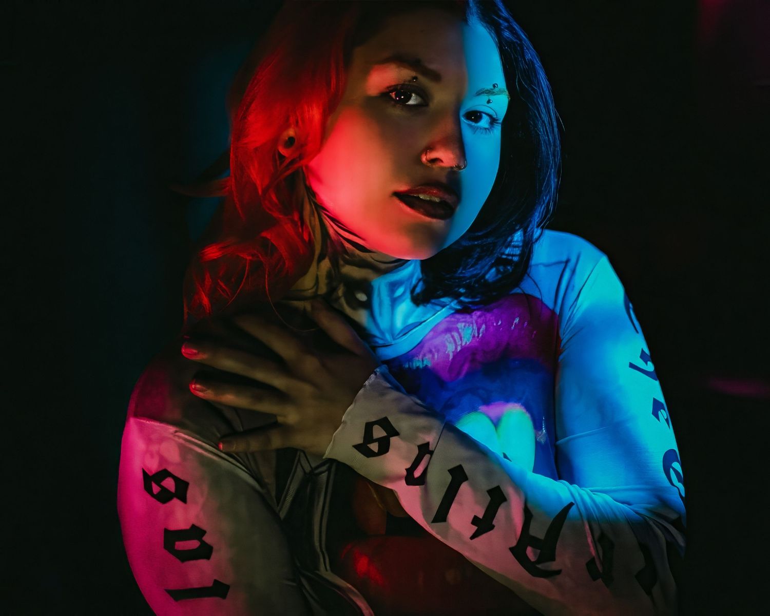 Woman with tattoos posed under dramatic red and blue neon lighting in a dark, moody artistic portrait photo.