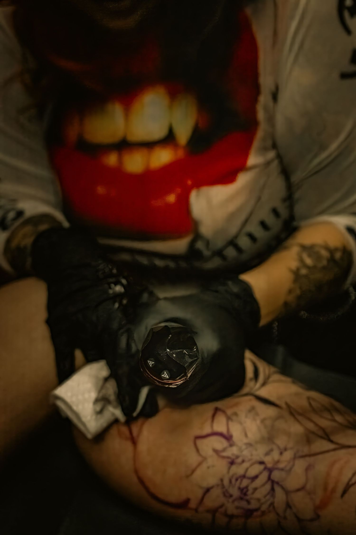 A tattoo artist wearing black gloves uses a tattoo machine to ink a floral design on a client's leg.