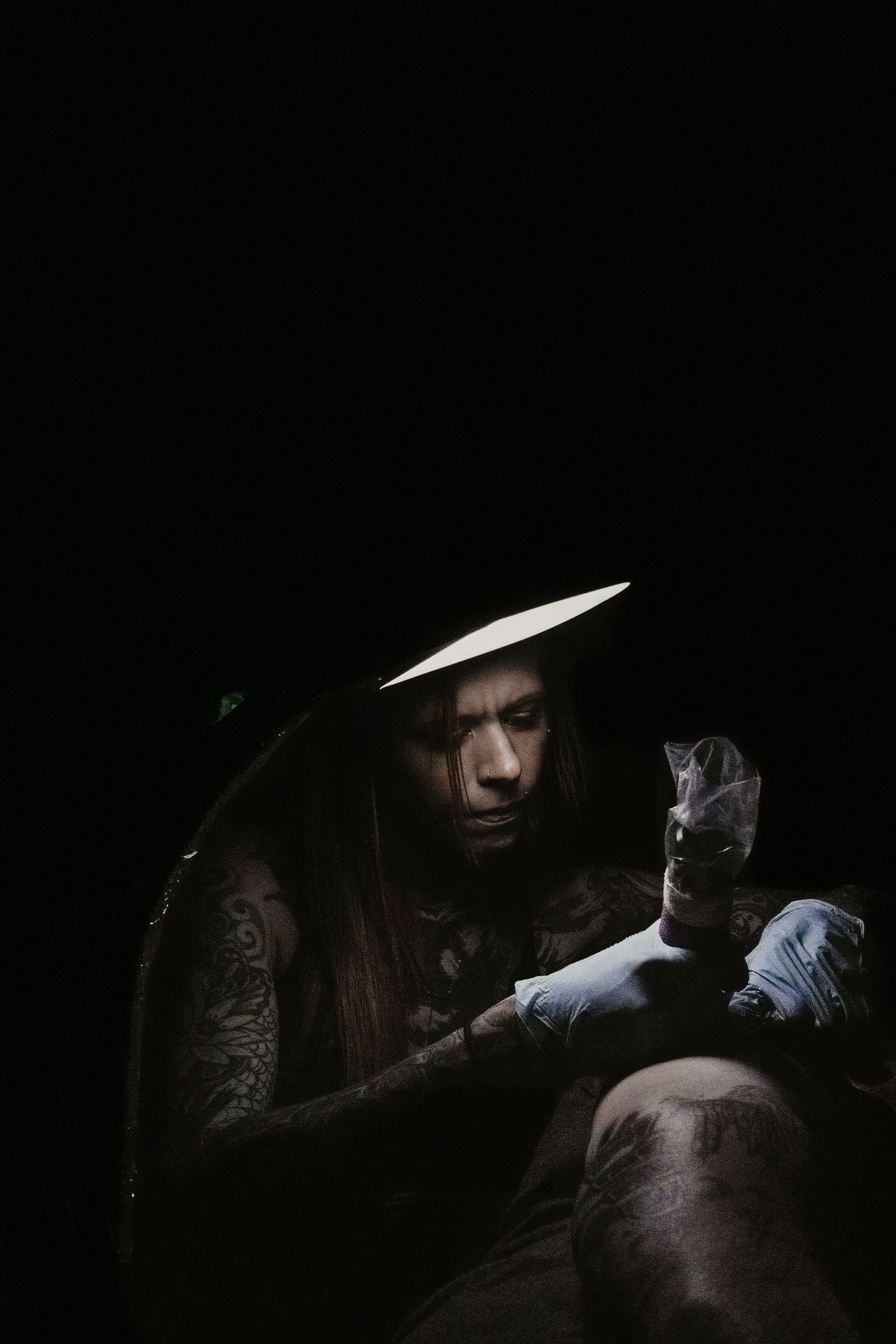 Female tattoo artist wearing a wide-brim hat and gloves works on a tattoo in a dimly lit studio.