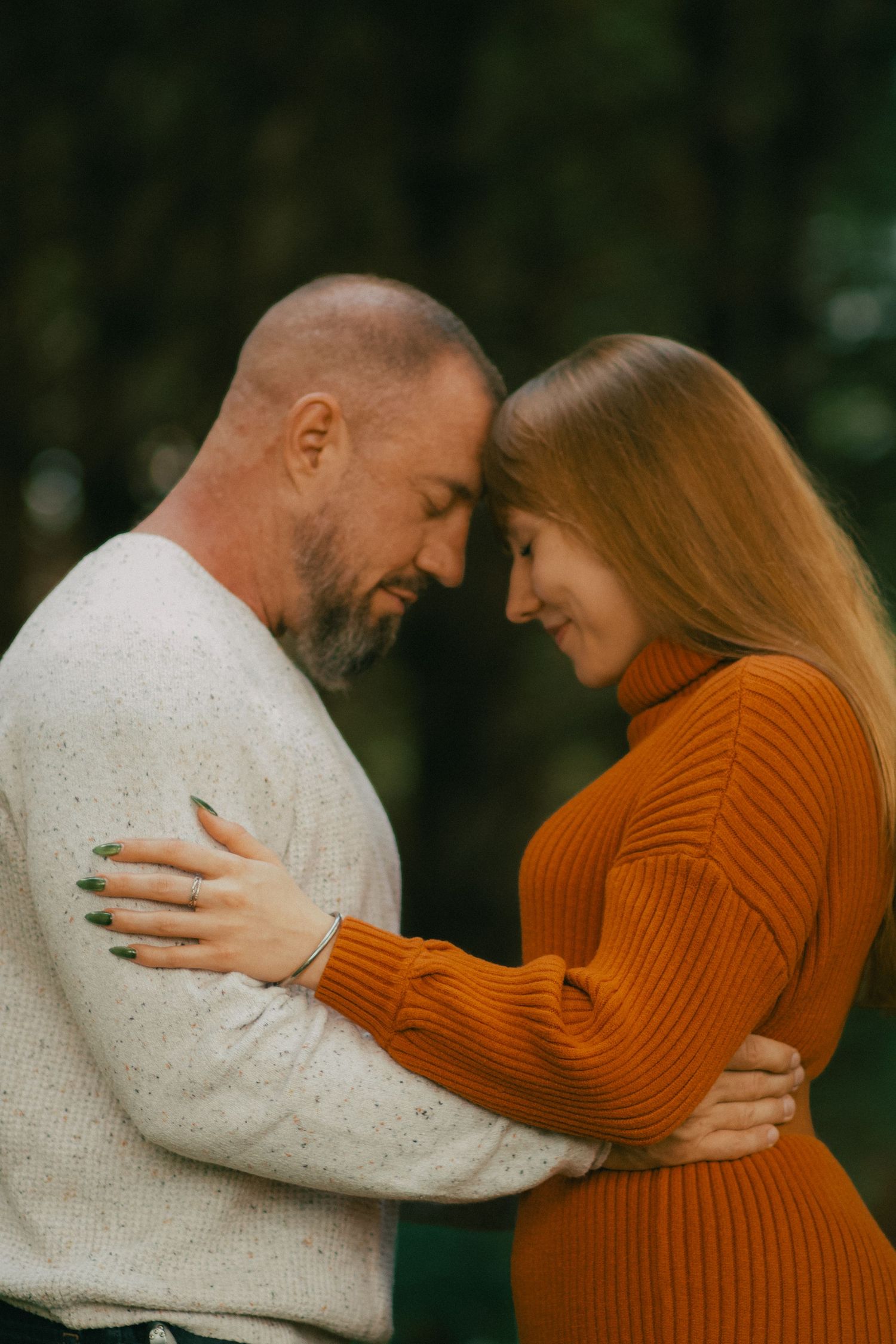 A couple in autumn sweaters embrace tenderly, touching foreheads in a romantic outdoor setting.