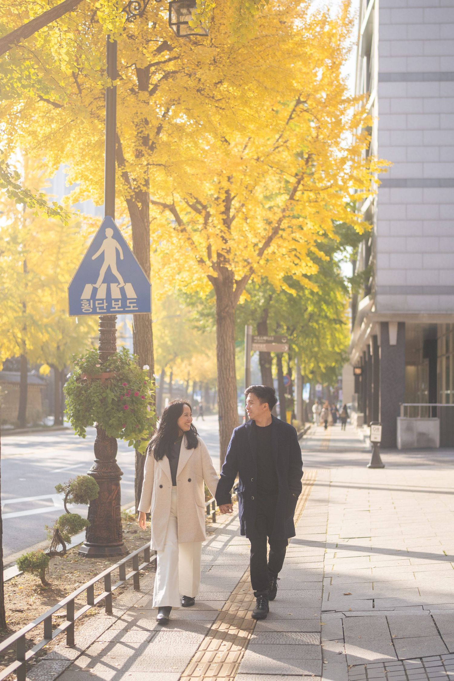 People stroll along a sidewalk under vibrant yellow autumn trees on a sunny day.